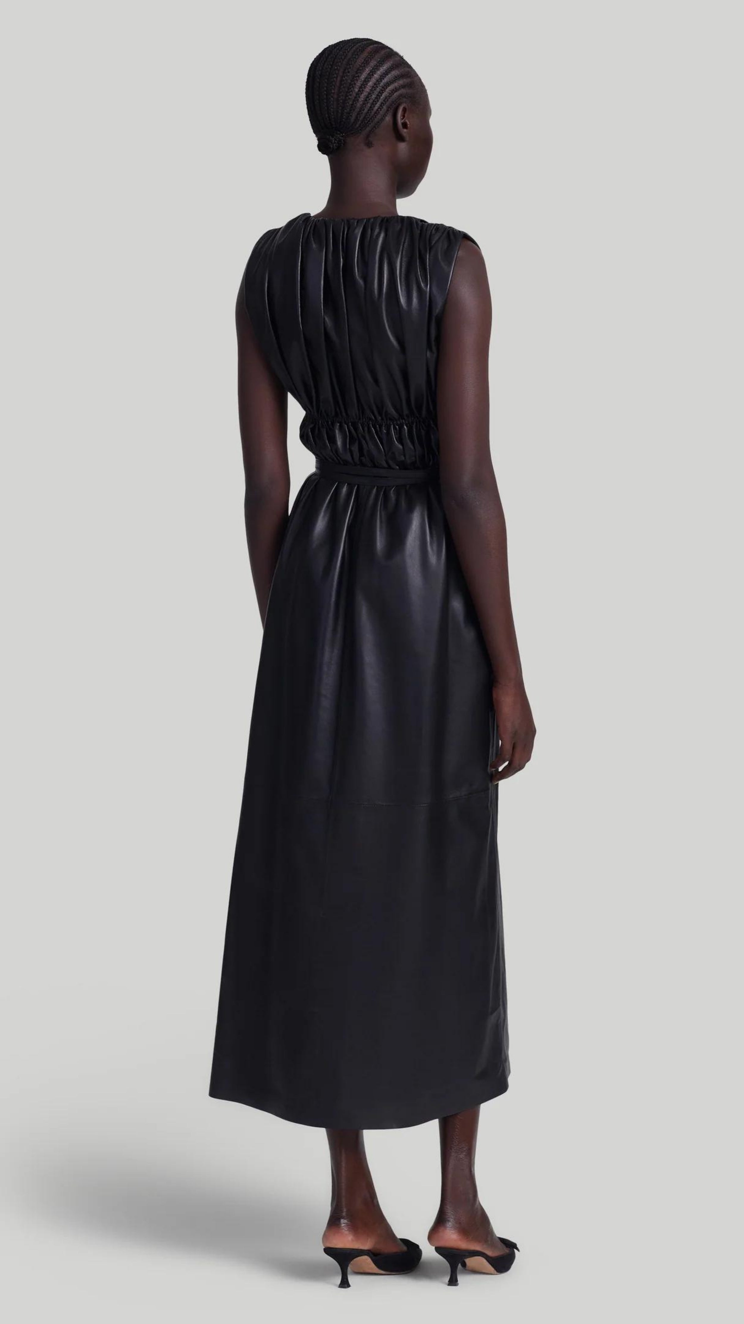 Altuzarra Fiona Dress, made from soft lamb leather. It features a maxi silhouette with a low V-neck, sleeveless, and ruching that folds at the waist with a thin leather belt. Shown on model facing back.