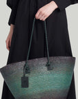 Altruzarra Large Watermill Bag in Campo. A summer raffia tote bag handmade in Mexico. It has a ombre tone with black and green. With a braided leather strap and black leather details. This photo shows the bag from the front view displayed by a model. Available at experience 27 in Madrid Spain. 