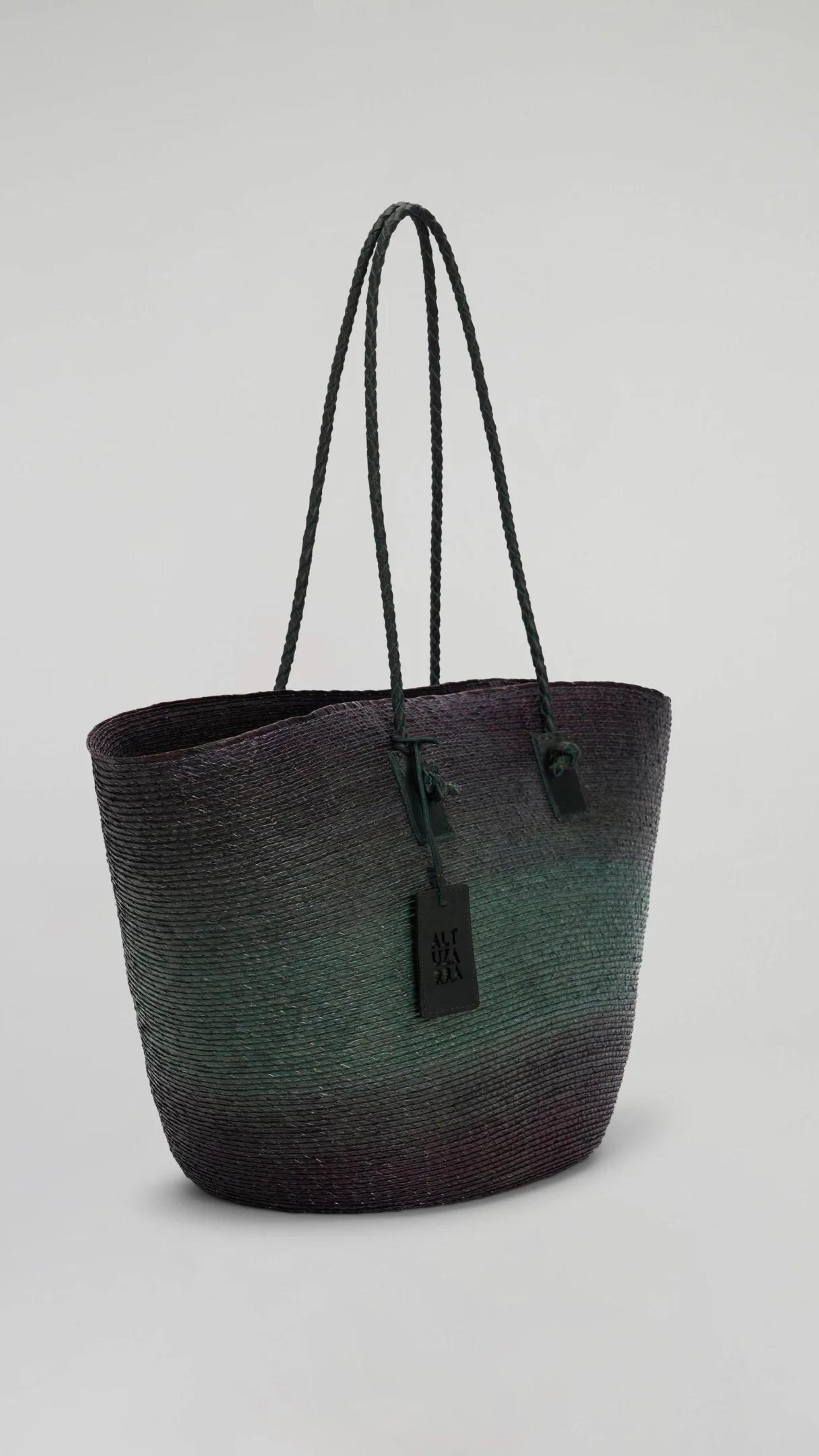 Altruzarra Large Watermill Bag in Campo. A summer raffia tote bag handmade in Mexico. It has a ombre tone with black and green. With a braided leather strap and black leather details. This photo shows the bag from the front side view. Available at experience 27 in Madrid Spain. 