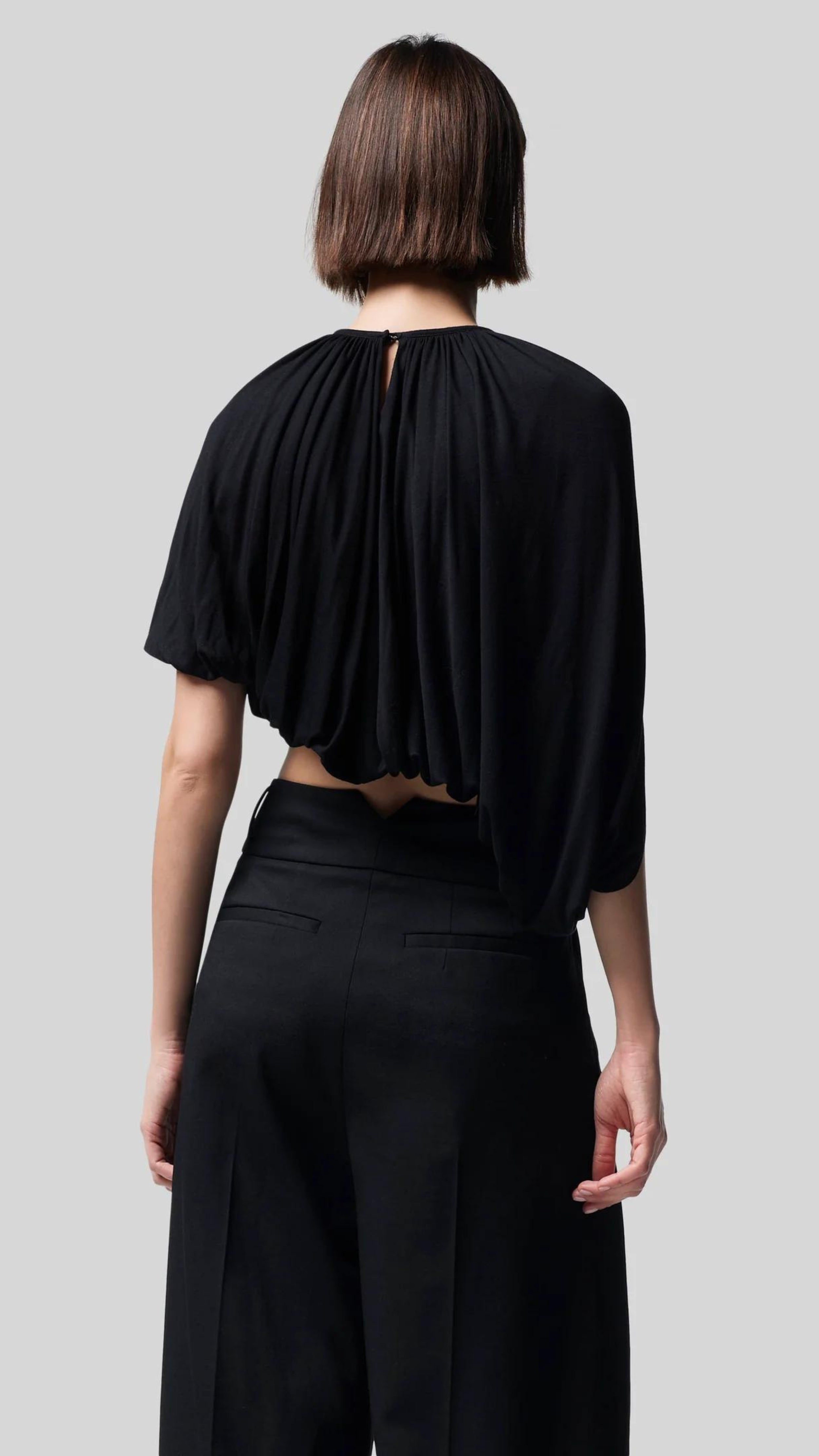 Altuzarra Naxos Top in Black. Draped jersey create a fluid look with an asymmetrical hemline. Shown on model facing to the back.