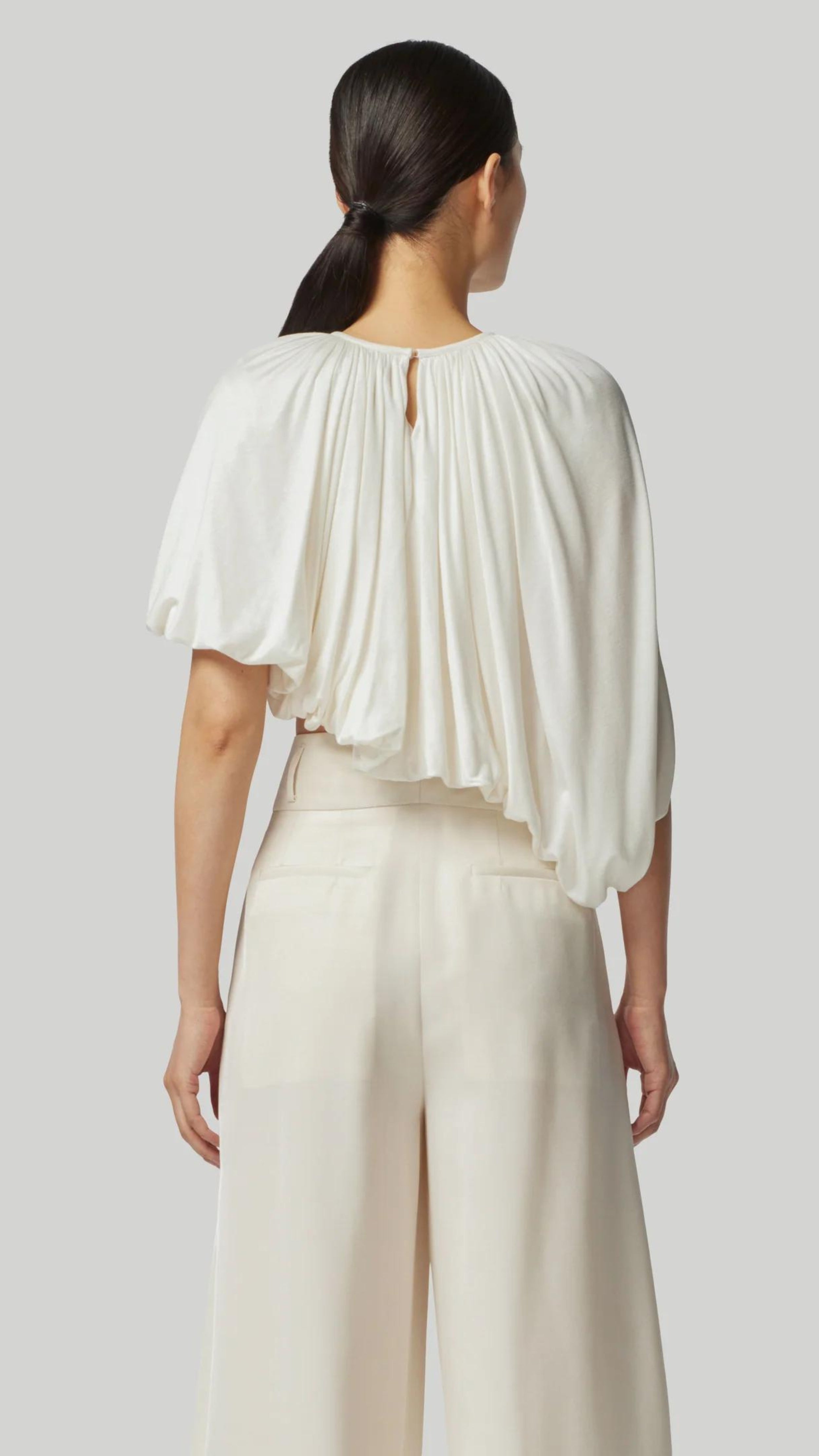 Altuzarra Naxos Top in Ivory. Draped jersey create a fluid look with an asymmetrical hemline. Shown on model facing to the back