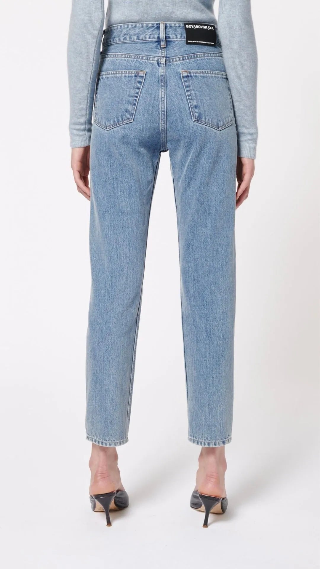 Boyarovskaya Slim Ring Jean. Light color stone denim with a slightly tapered leg and falls to just above the ankle. Zipper closure with silver ring detail on the front right. Product shown on model facing back.