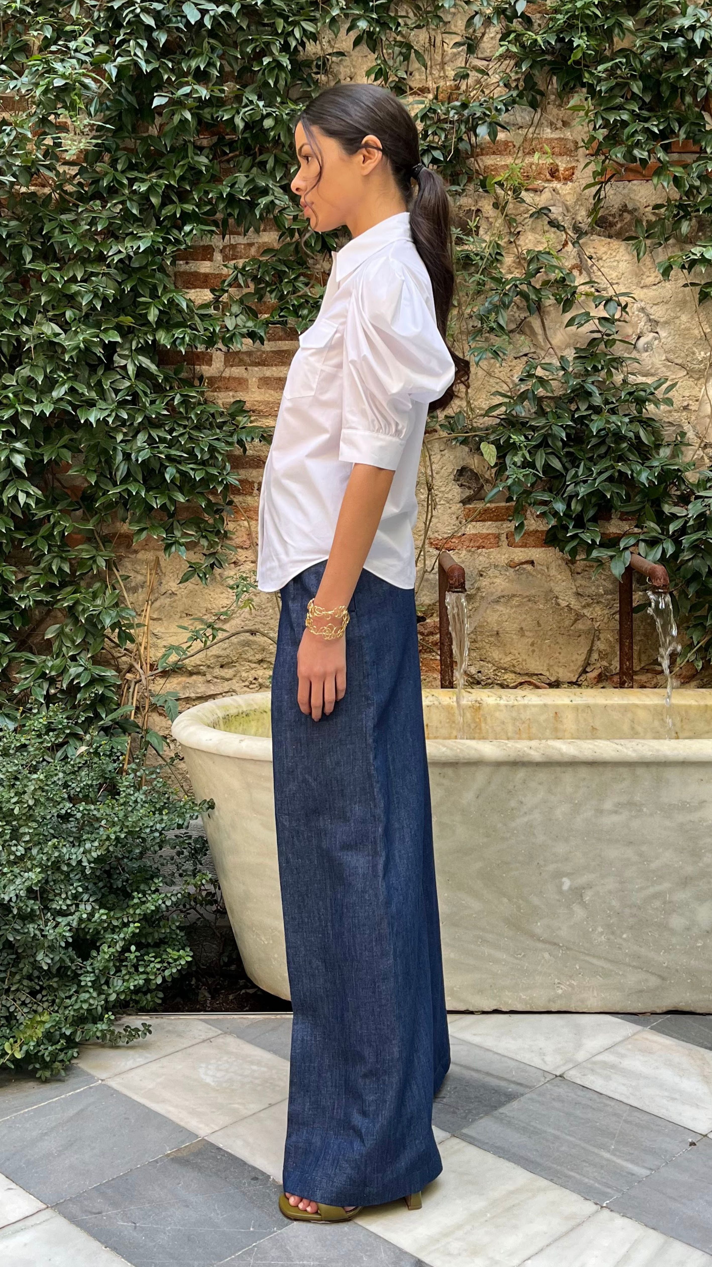Rochas Paris Cotton Poplin Short Sleeved Shirt. A button up blouse in lightweight cotton it has two front pockets and puffed sleeves. Photo shown on model also wearing Rochas wide leg denim trousers from a side view.