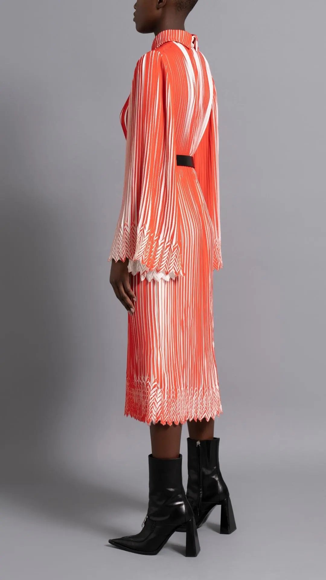 Thebe Magugu Chevron Pleated Dress. Stunning tomato red and white pleated dress with a collar, fitted waist and double chevron hem at the sleeves. It features a hidden zipper in the back. Shown on model facing to the side and back.