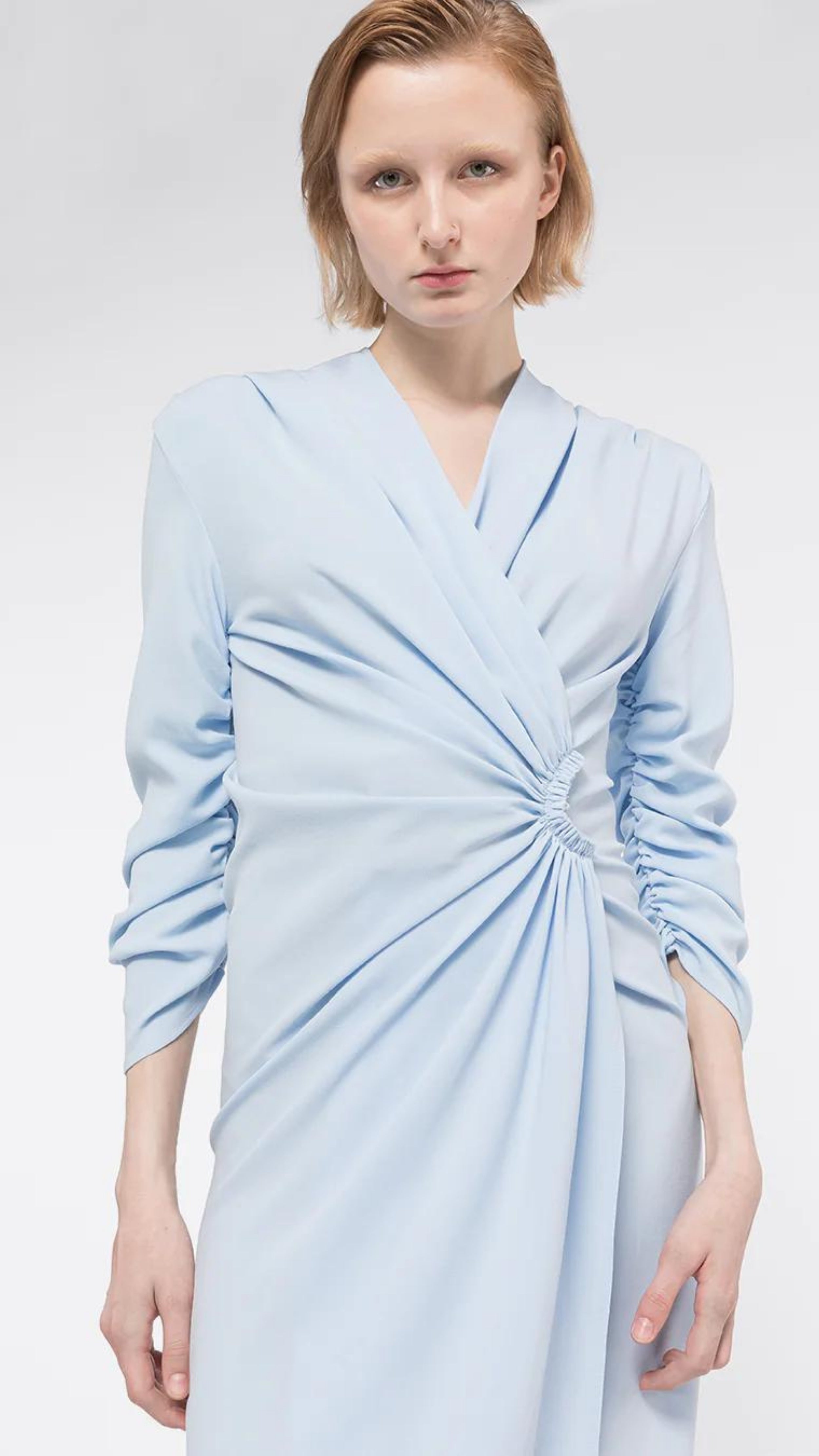 AZ Factory Lutz Huelle, Ella Dress. Crafted with a crepe texture in sky blue hue. V neck and draped, off center gathered front. Half length ruched sleeves  with a midi length skirt. Shown on model facing front, detail.