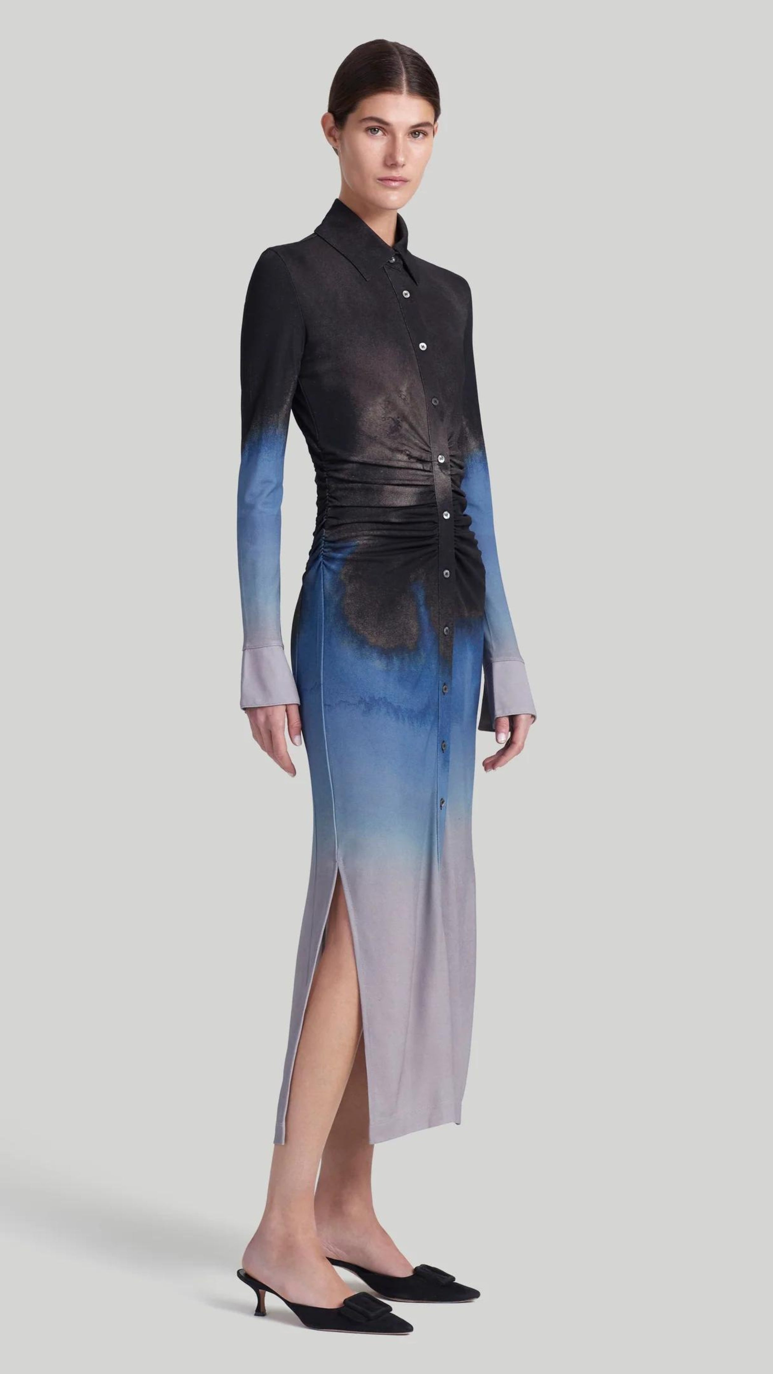 Altuzarra Claudia Dress in Eventide Made from jersey fabric, it is a slim-fitting midi dress featuring ruched detail at the waist, shirt dress buttons up the front, and side slits at the hem. The color is an ombre of deep grey, blue, and pale grey. Shown on model facing front side.