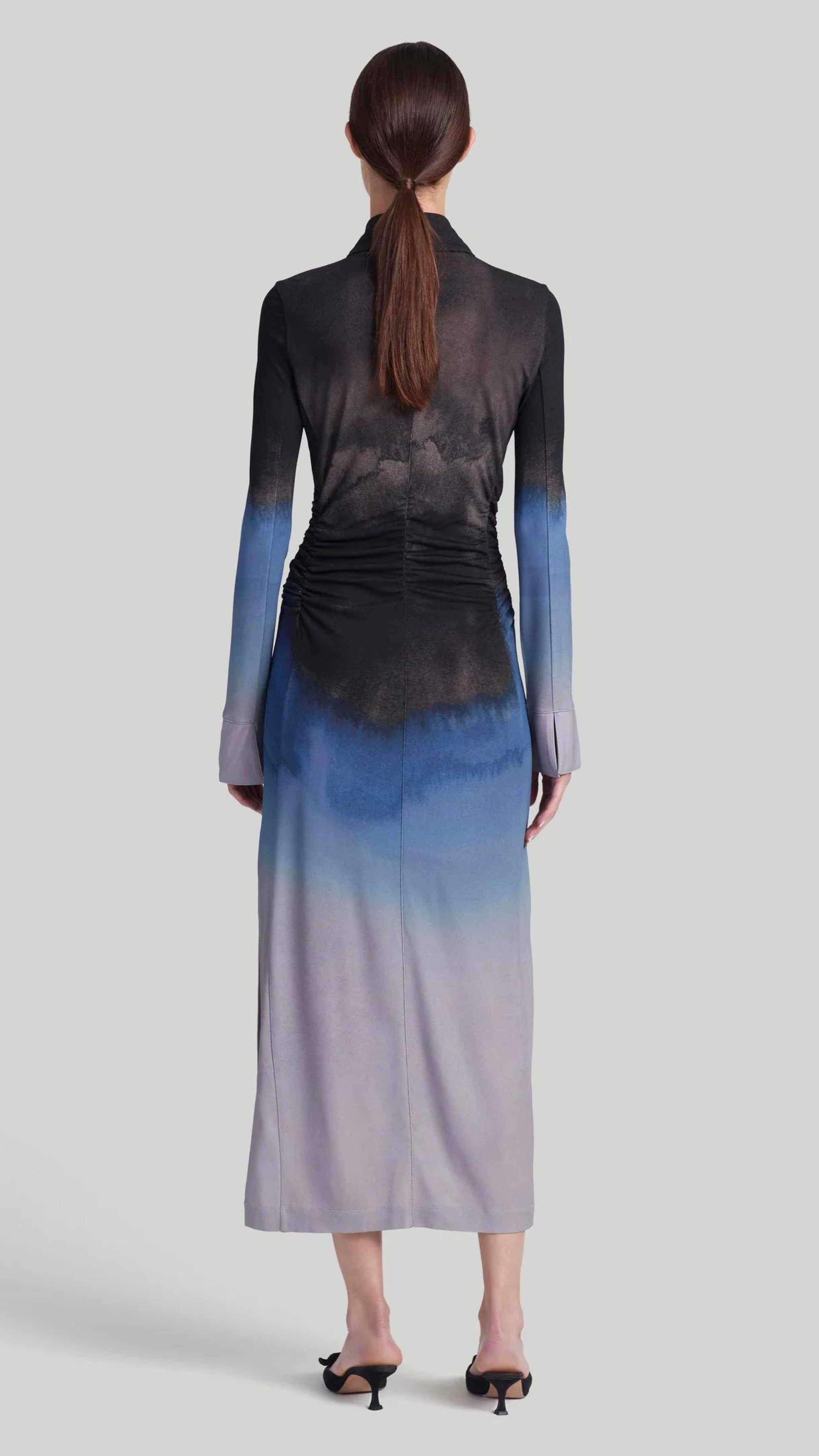 Altuzarra Claudia Dress in Eventide Made from jersey fabric, it is a slim-fitting midi dress featuring ruched detail at the waist, shirt dress buttons up the front, and side slits at the hem. The color is an ombre of deep grey, blue, and pale grey. Shown on model facing back.