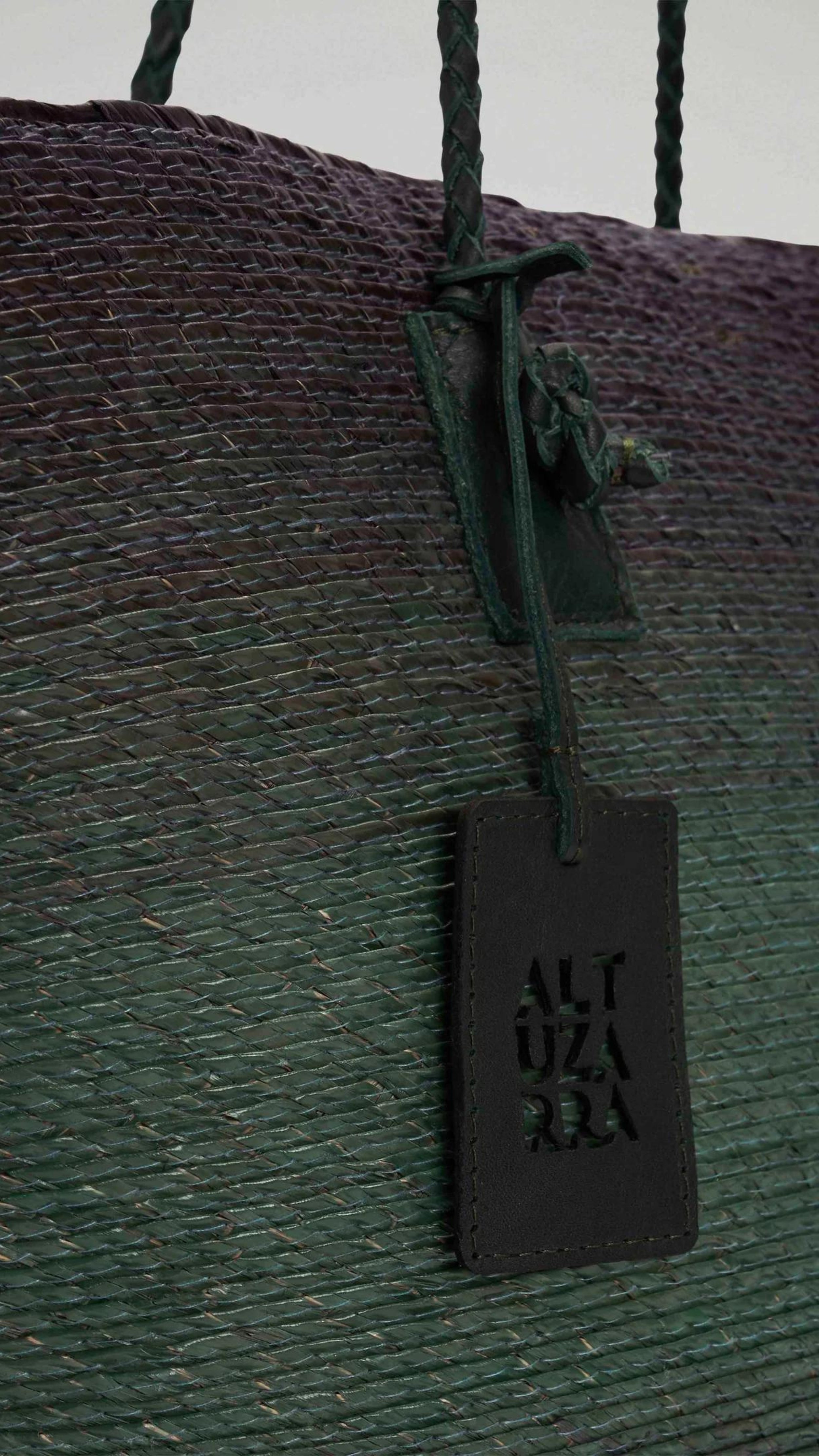 Altruzarra Large Watermill Bag in Campo. A summer raffia tote bag handmade in Mexico. It has a ombre tone with black and green. With a braided leather strap and black leather details. This photo shows a close up of the Altruzarra leather tag.. Available at experience 27 in Madrid Spain. 