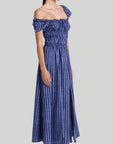 Altuzarra Lily Dress The 'Lily' dressmade from soft cotton. The midi dress  features a smocked construction and off-the-shoulder design. It has hidden pockets and a side slit in the skirt. In tones of blue. Shown on model facing side.