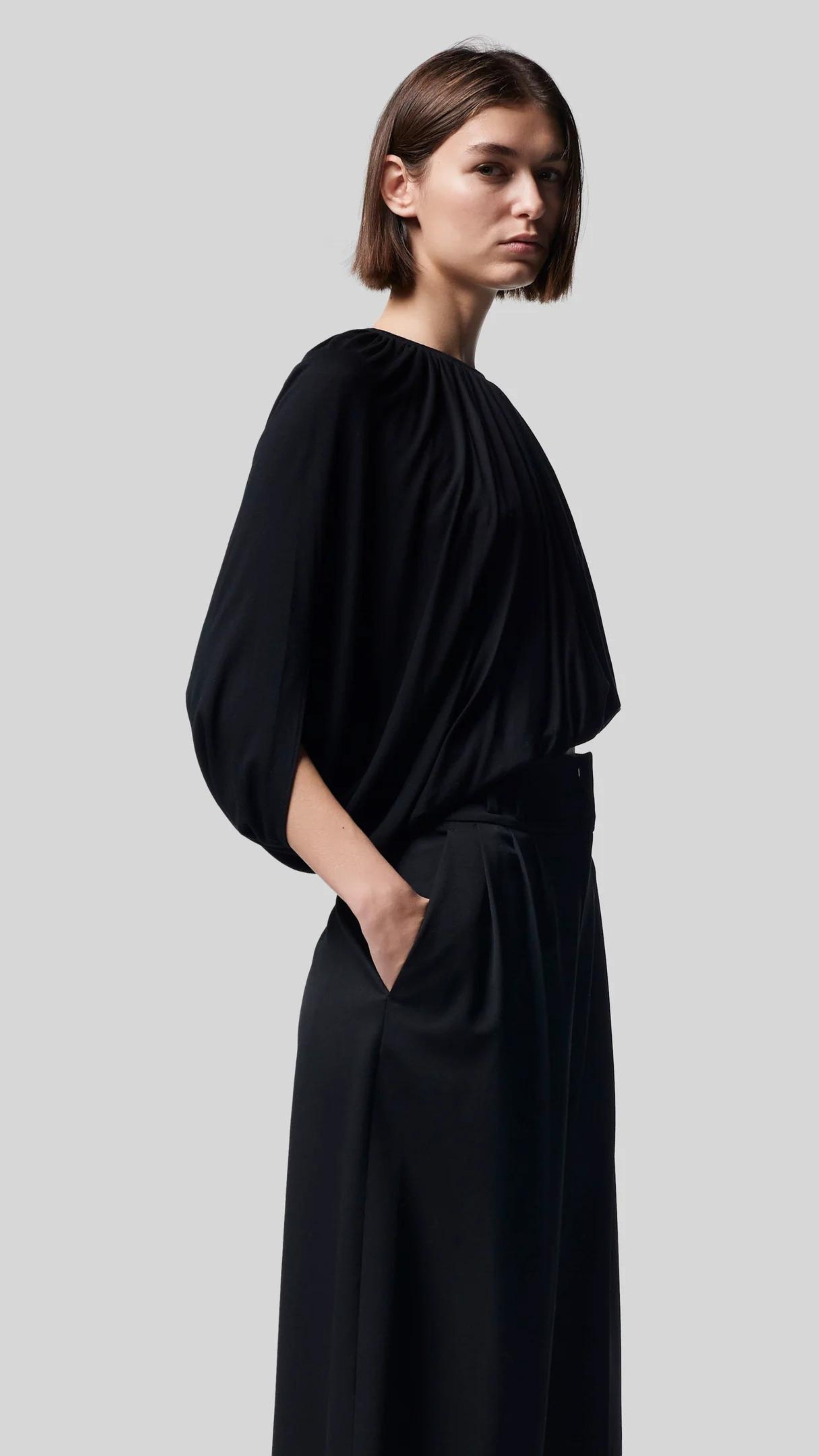 Altuzarra Naxos Top in Black. Draped jersey create a fluid look with an asymmetrical hemline. Shown on model facing to the side.