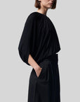 Altuzarra Naxos Top in Black. Draped jersey create a fluid look with an asymmetrical hemline. Shown on model facing to the side.