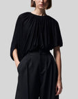 Altuzarra Naxos Top in Black. Draped jersey create a fluid look with an asymmetrical hemline. Shown on model facing to the front.