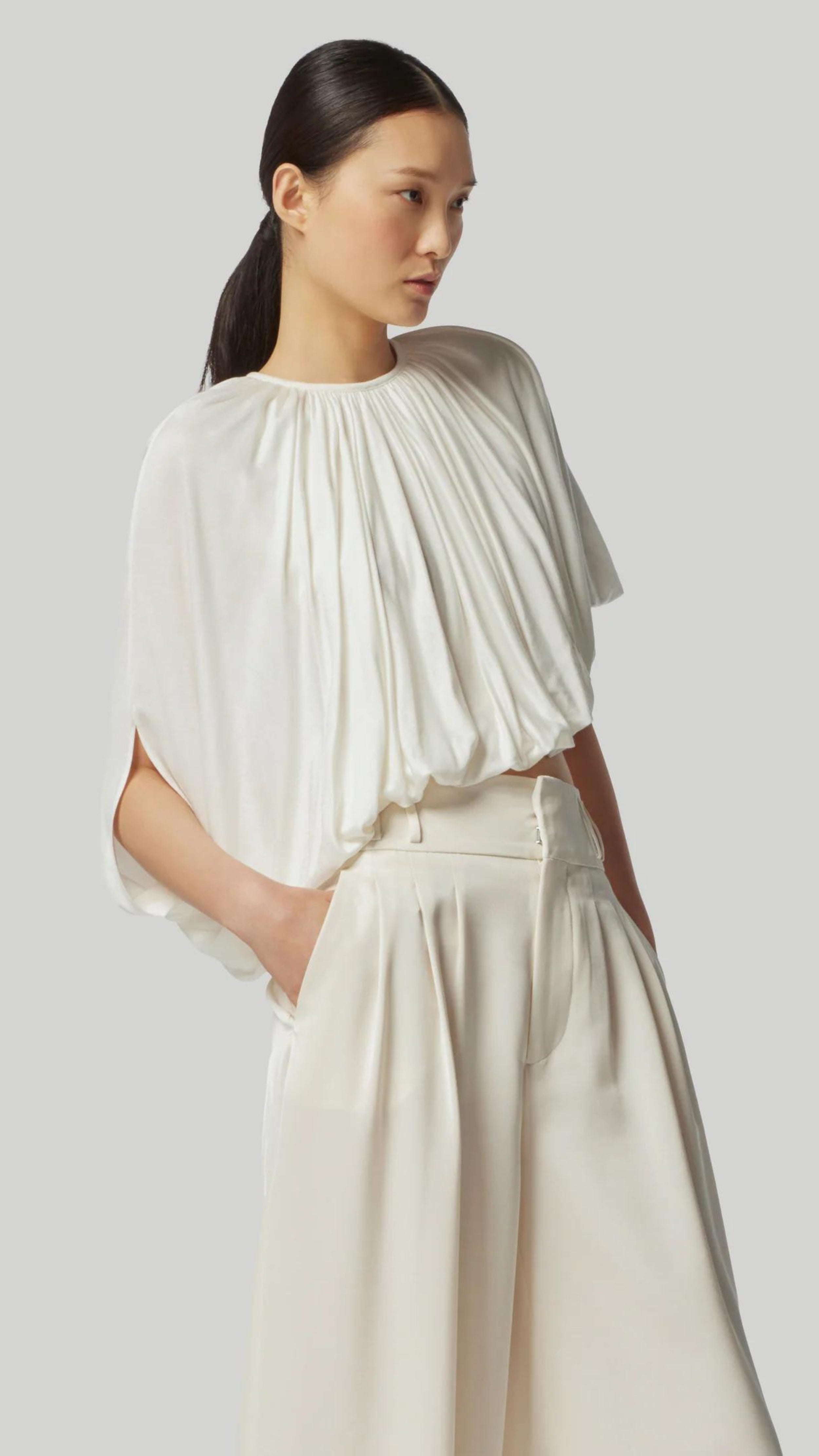Altuzarra Naxos Top in Ivory. Draped jersey create a fluid look with an asymmetrical hemline. Shown on model facing to the front side.