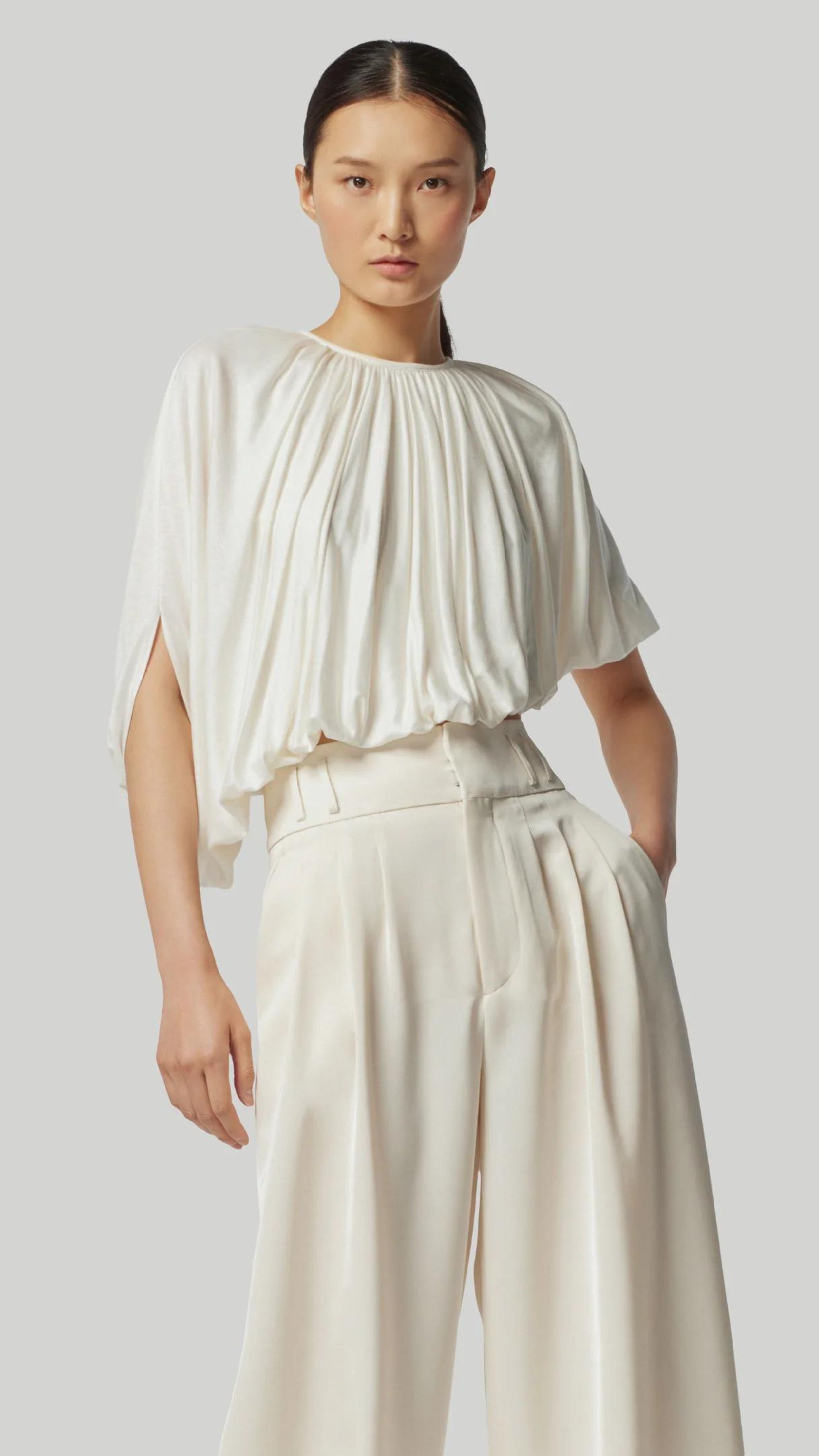 Altuzarra Naxos Top in Ivory. Draped jersey create a fluid look with an asymmetrical hemline. Shown on model facing to the front.