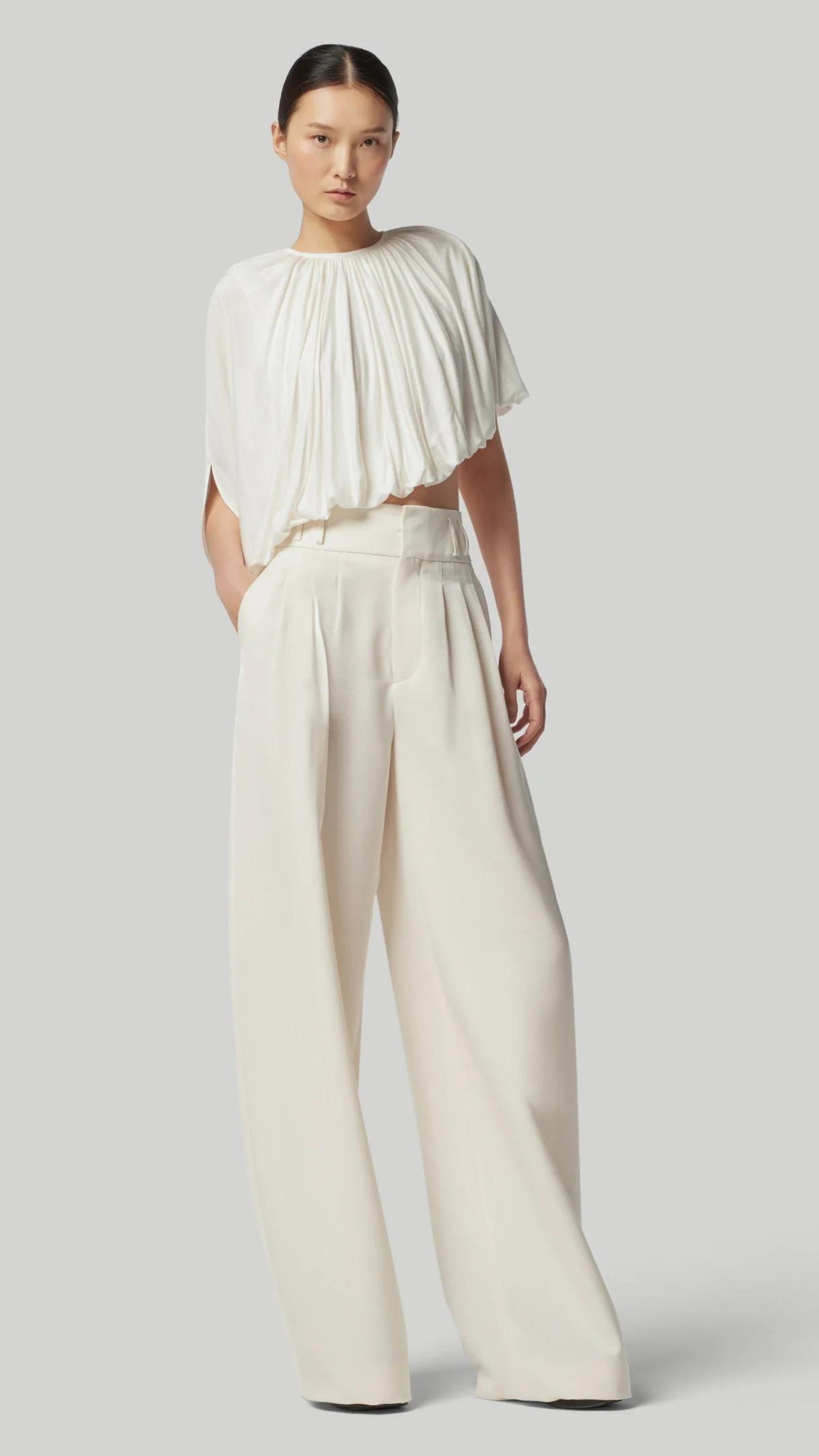 Altuzarra Naxos Top in Ivory. Draped jersey create a fluid look with an asymmetrical hemline. Shown on model facing to the front side.