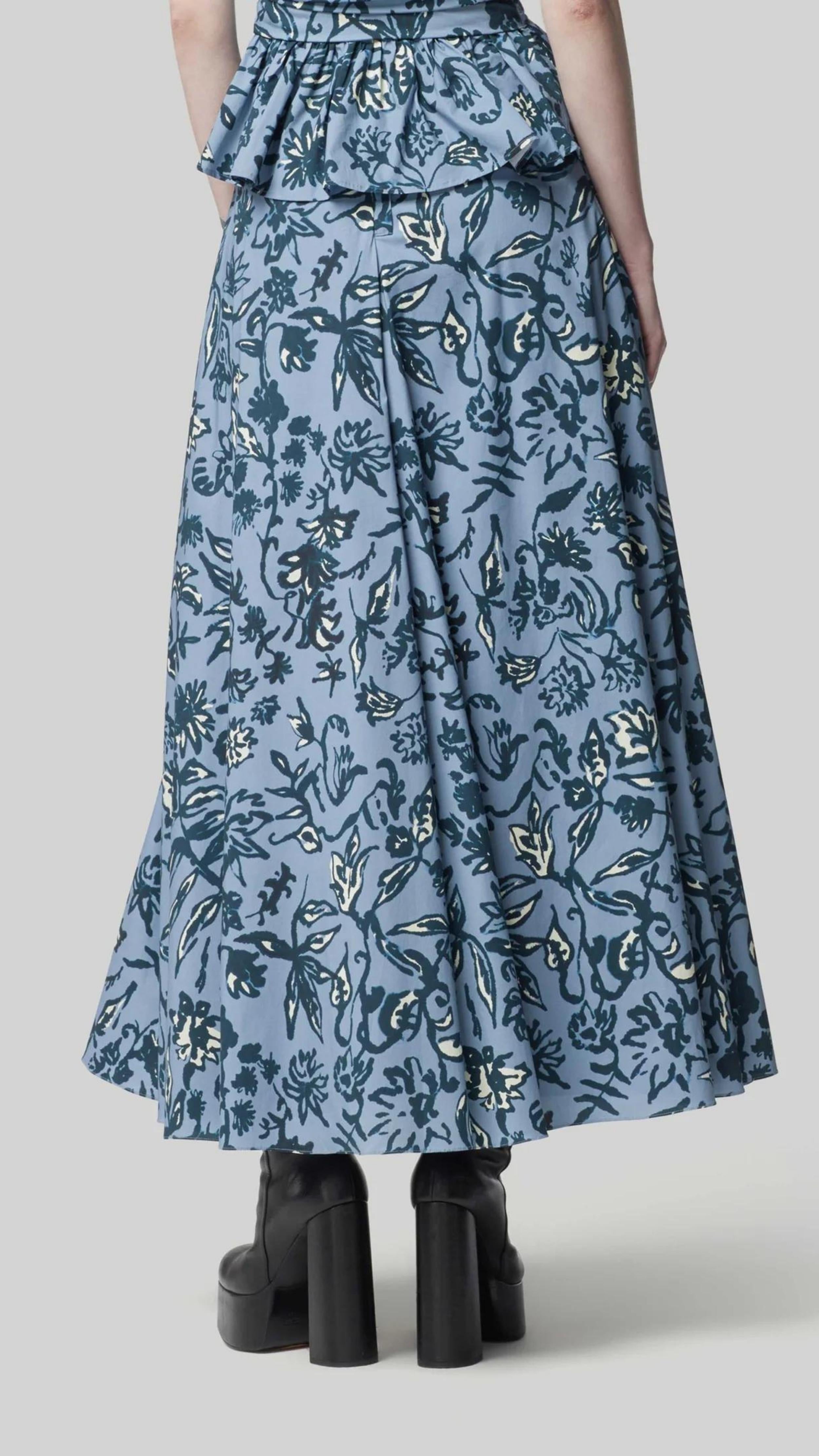 Altuzarra Pythia Skirt in Amsonia Shibori Flower. A midi length, aline style skirt with beautiful twist detail at the waist. Crafted in 100% cotton with a natural ecru color floral print over a light blue background. Shown on model facing back. Available at experience 27 in Madrid spain.