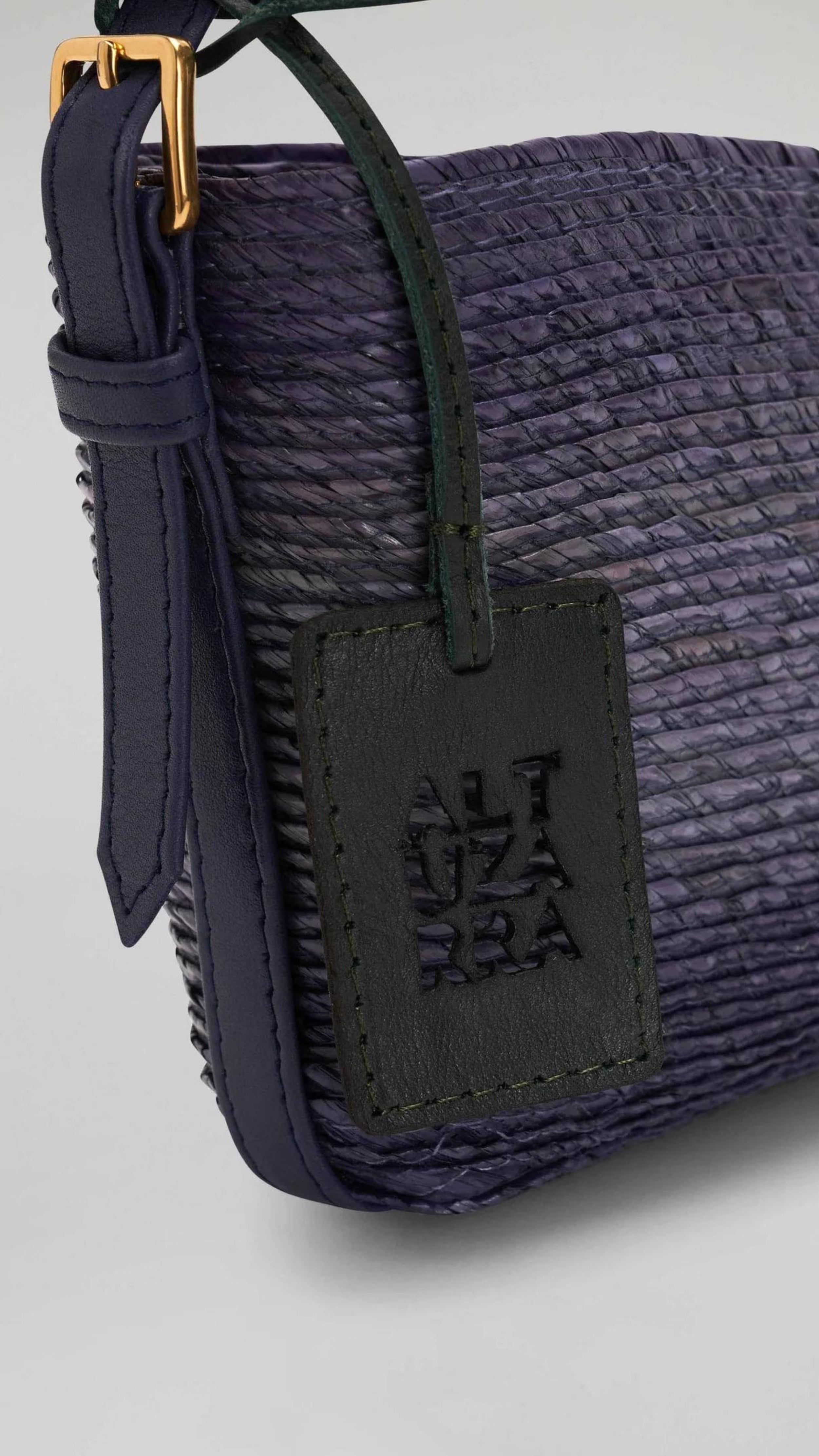 Altruzarra WATERMILL SHOULDER BAG in Murex. Raffia summer purse in deep blue tones and navy blue leather detailing and gold hardware. Adjustable strap for shoulder or long length. Close up view of the Altruzarra blue leather tag.. Available at Experience 27 Madrid Spain.