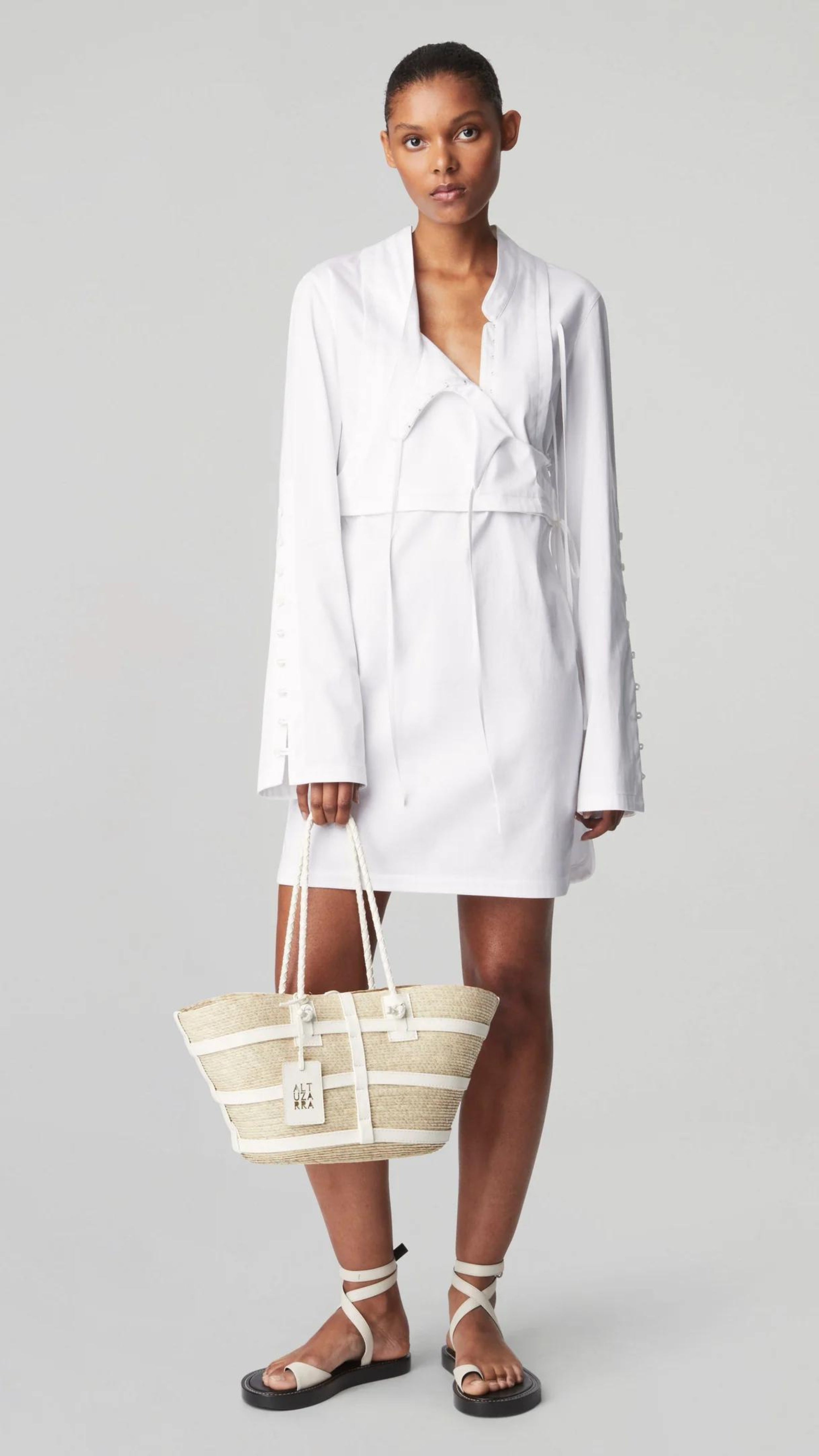 Altuzara Watermill Bag Small in White. Crafted in Mexico from natural palm fibers and detailed with soft white leather. The hand straps are braided leather and it comes with a laser cut detailed Altuzarra hang tag. This photo shows the bag being held by the model.