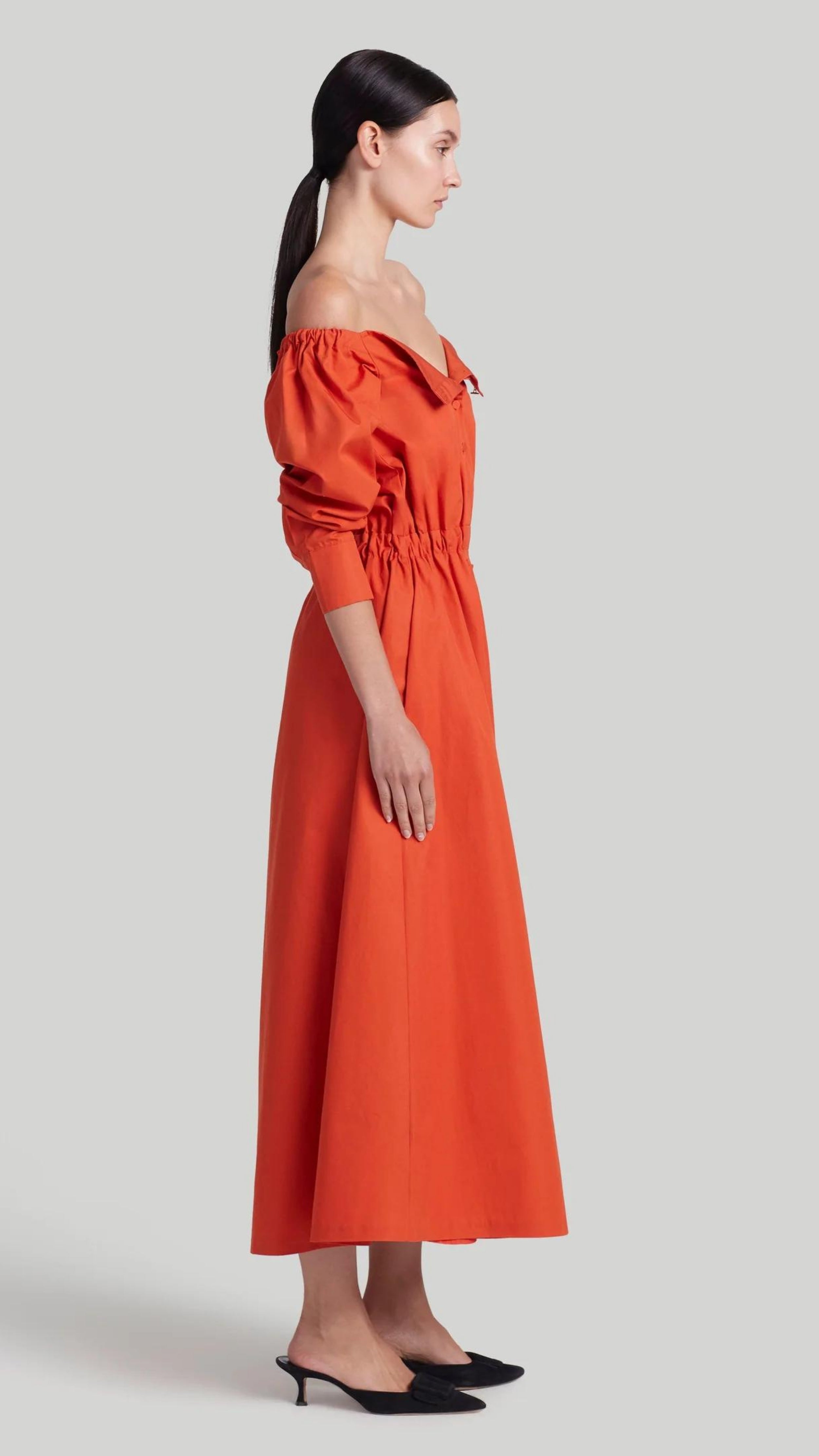 Altuzarra Zora dress shirt dress style soft cotton midi dress in red orange color. Made in Italy. The dress has off the shoulder neckline, puff sleeves and an elasticated waist waistband. Shown on model facing side