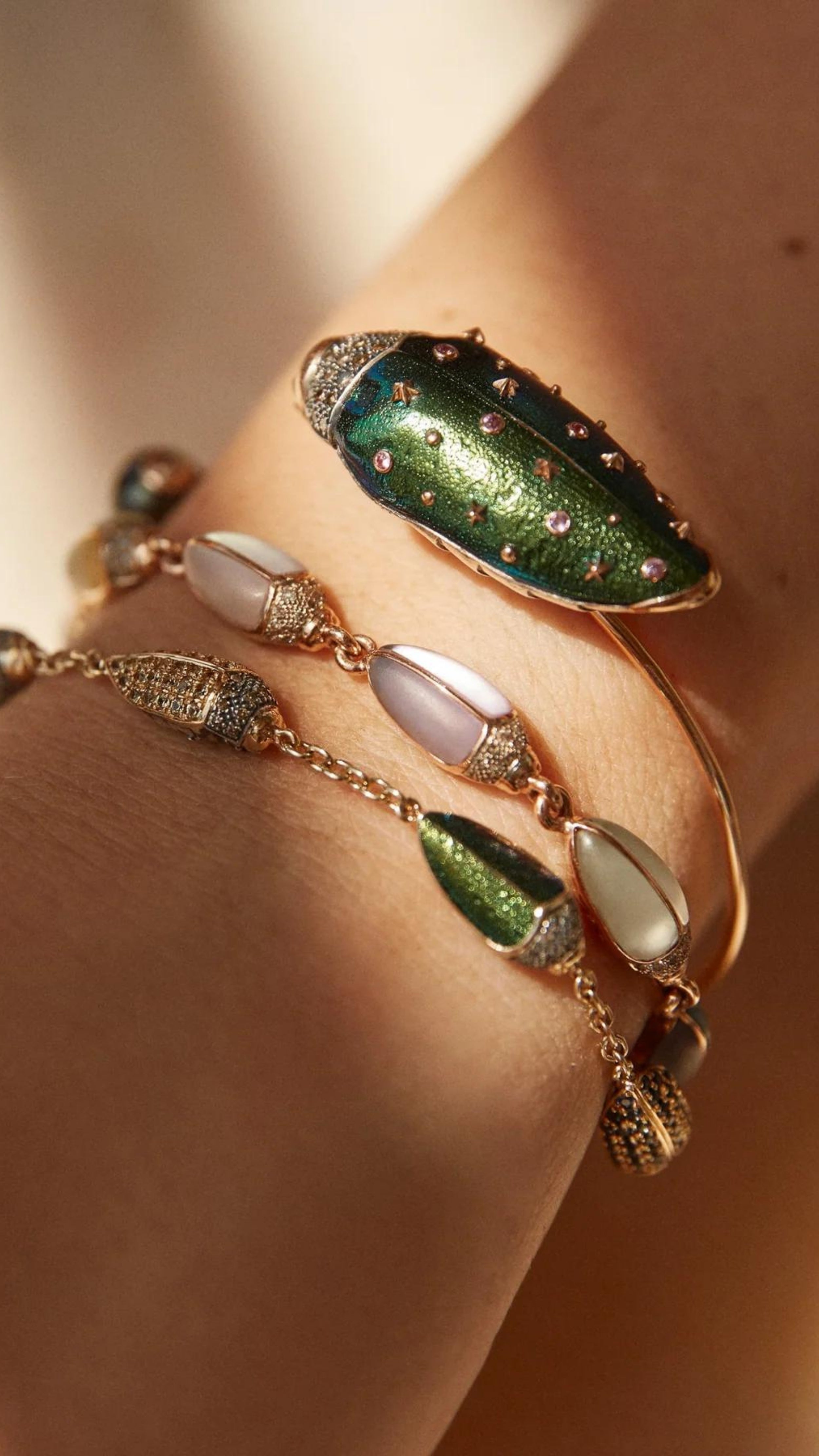 Bibi van der Velden Mini Scarab Eternity Bracelet. Made in 18k rose gold, this bracelet has nine  scarab beetles, each made with translucent, matte gemstones in soft shades. The beetles&#39; heads are paved with pale brown diamonds. Shown being worn on an arm.