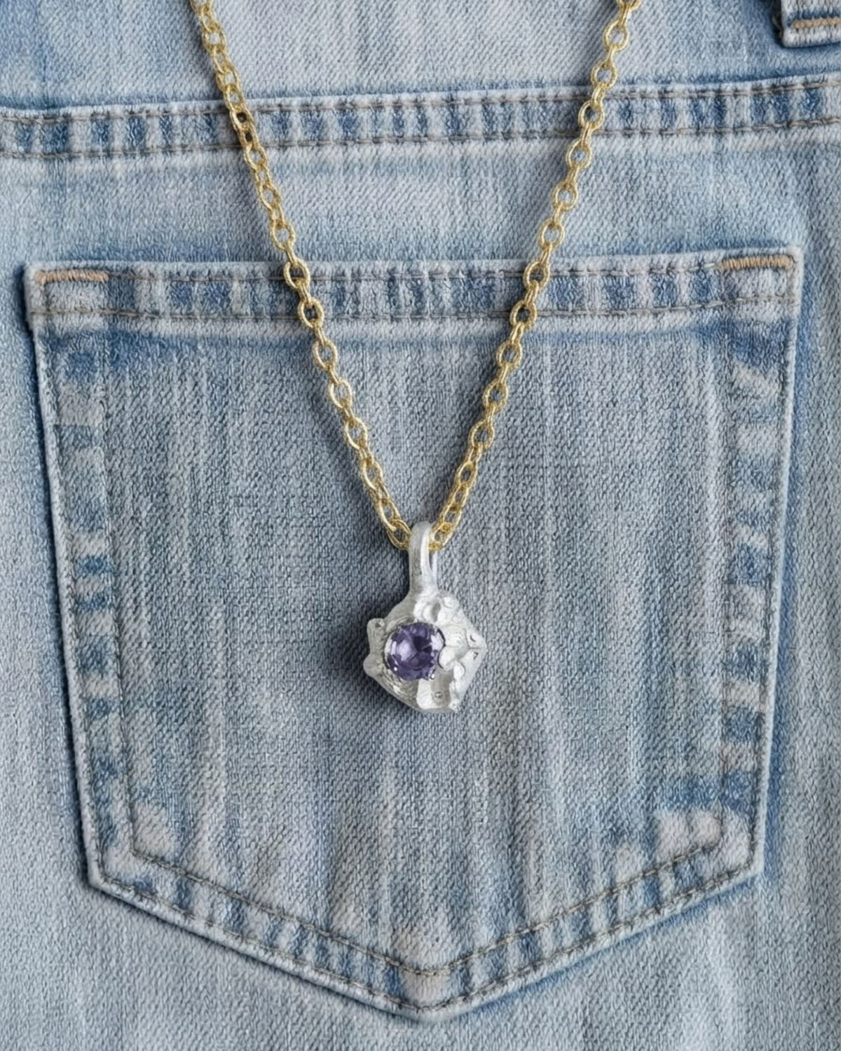 Gold chain necklace with a sterling silver amethyst pendant draped over a denim pocket