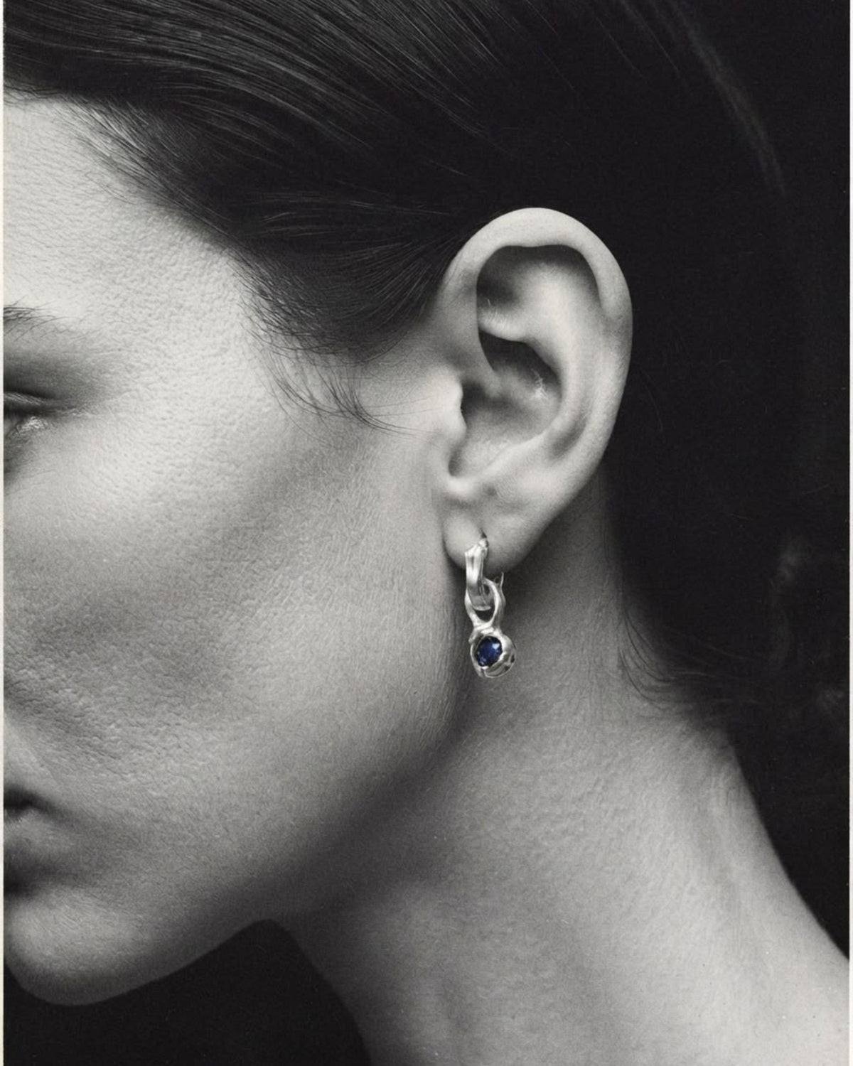 1. Close-up of a sterling silver hoop earring with a blue topaz drop, worn on a model's ear, black and white
