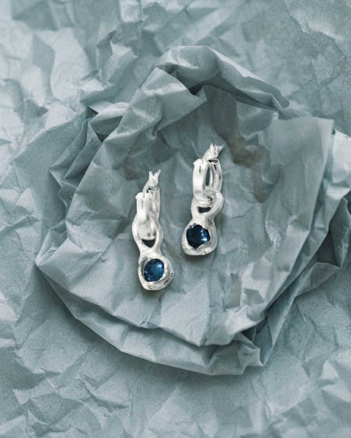 Two sterling silver hoop earrings with blue topaz drops resting on grey textured paper