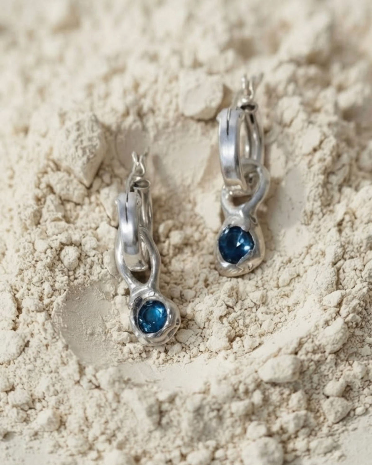 Sterling silver hoop earrings with blue topaz drops resting on beige textured surface