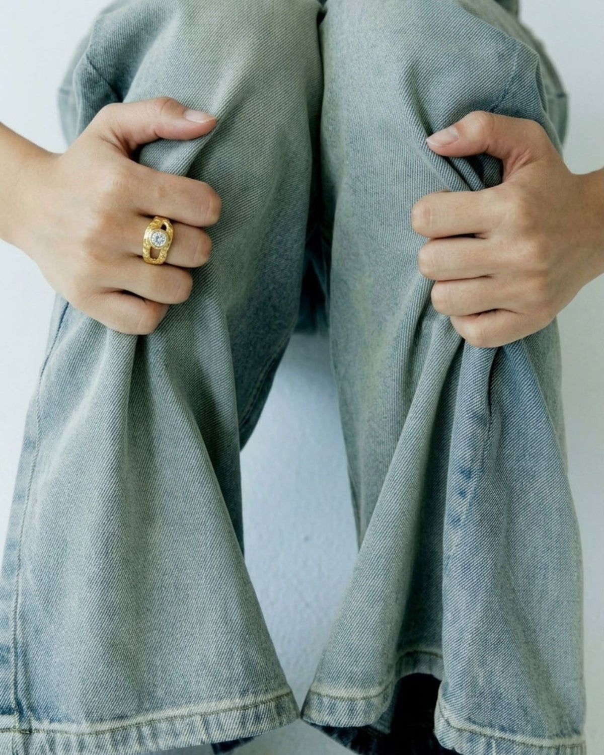 Close-up of an 18k gold vermeil ring with a round white stone, worn on a model's hand gripping denim fabric