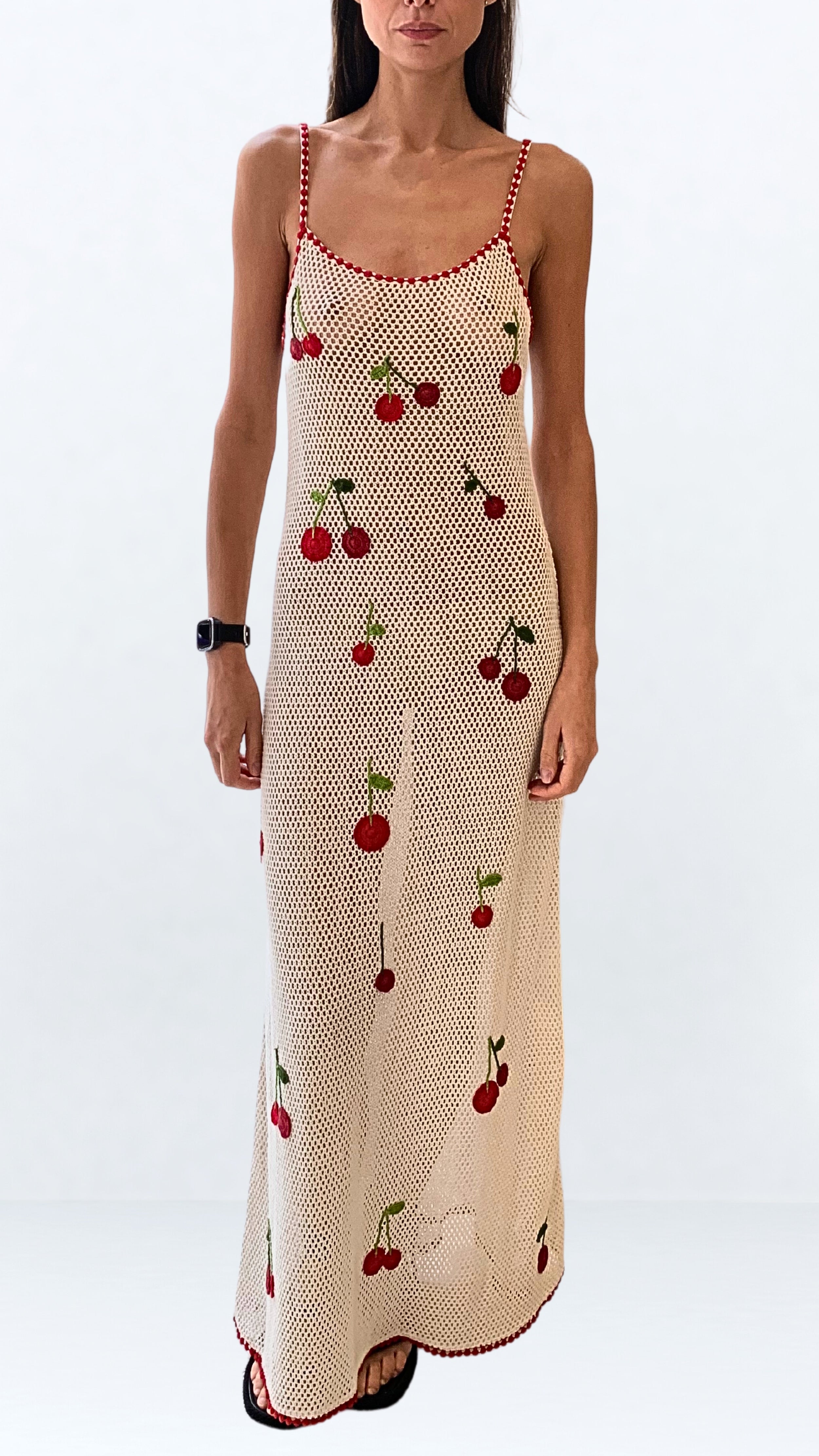 Leslie Amon Treasure Cherry Dress Maxi in White. Crochet knit cotton dress with playful cherry embroidery. Spaghetti strap sleeves in a red ribbon. Maxi length. Shown on model facing front.