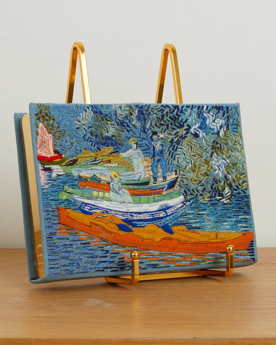 Lifestyle shot of the Bank of the Oise at Auvers clutch propped on a gold easel stand, showing the embroidered front panel at an angle on a wooden surface.