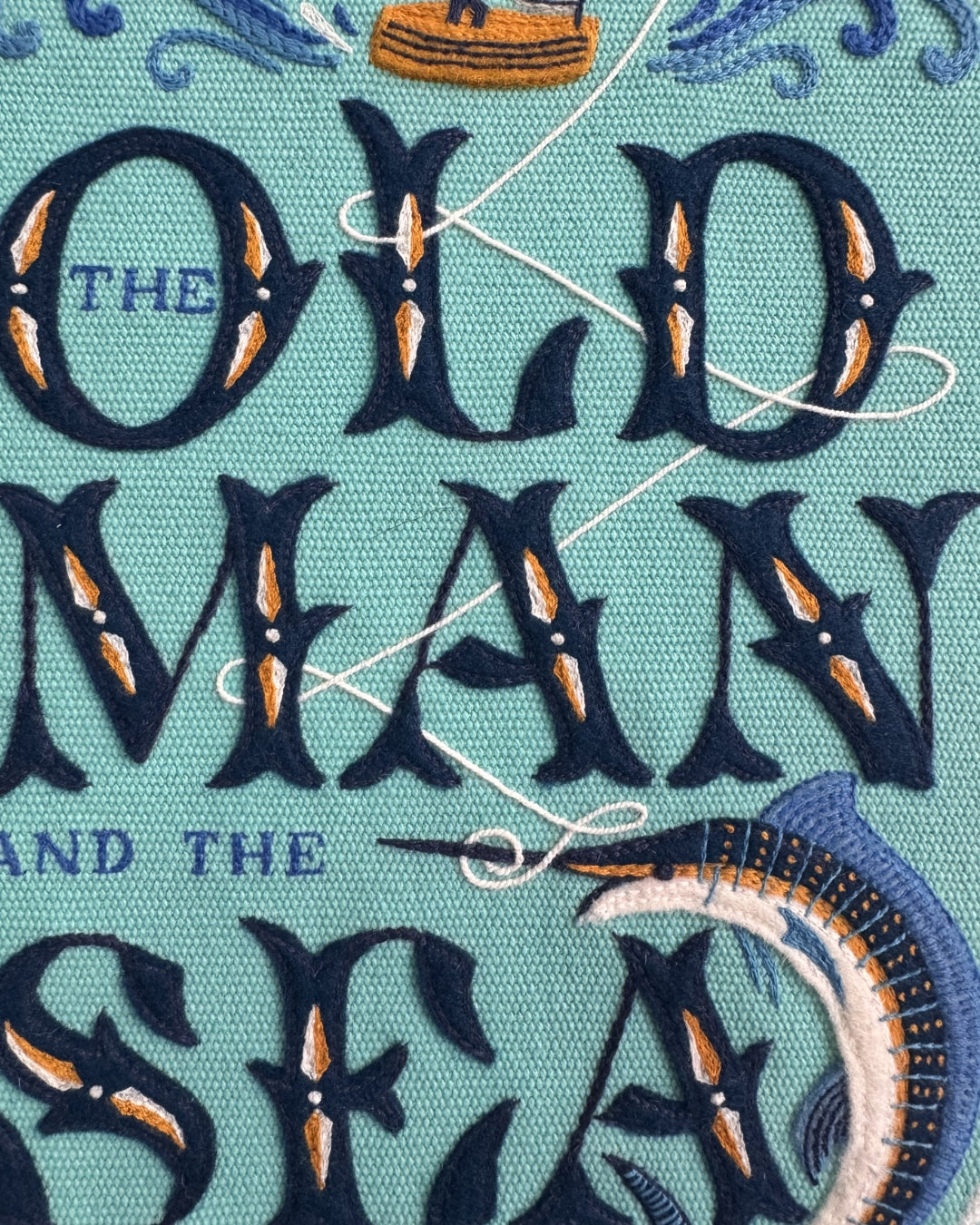 Close-up detail of the hand embroidery on the Old Man and the Sea clutch, showing the felt appliqué lettering, silk thread marlin, fishing line and boat motifs on the teal cotton ground.