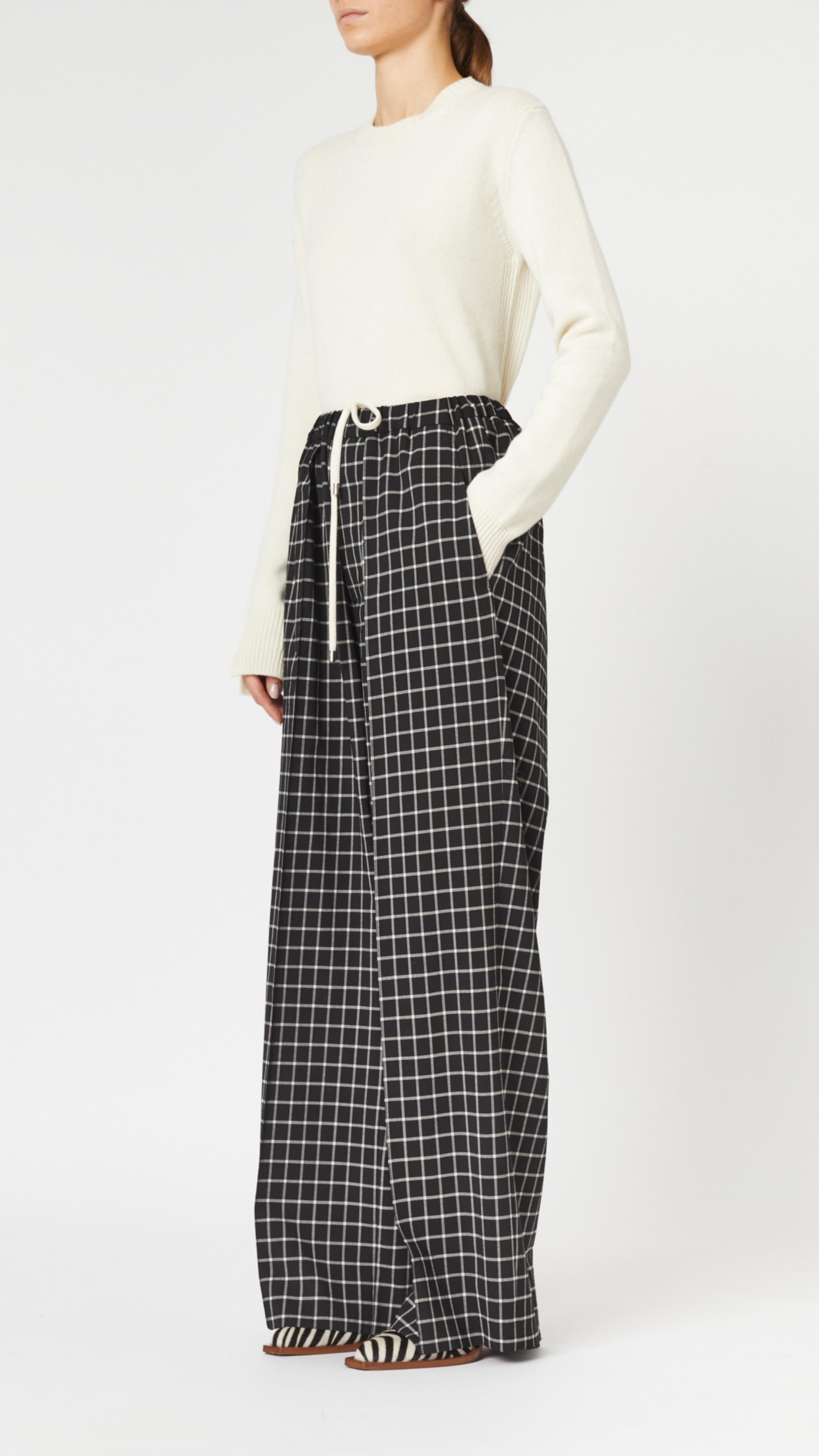 Plan C Checked Black Trousers. Loose fitting suit trousers with a drawstring adjustable waist in a classic black and white check patttern. Modern and oversized style. Shown on model facing to the side and with her hand in the pocket..