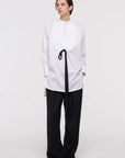 Plan C White Sartorial Poplin Shirt. Classic with a twist long white poplin blouse with front buttons. An added black ribbon detail in the front. Oversized cuffs at the long sleeves. Shown on model.