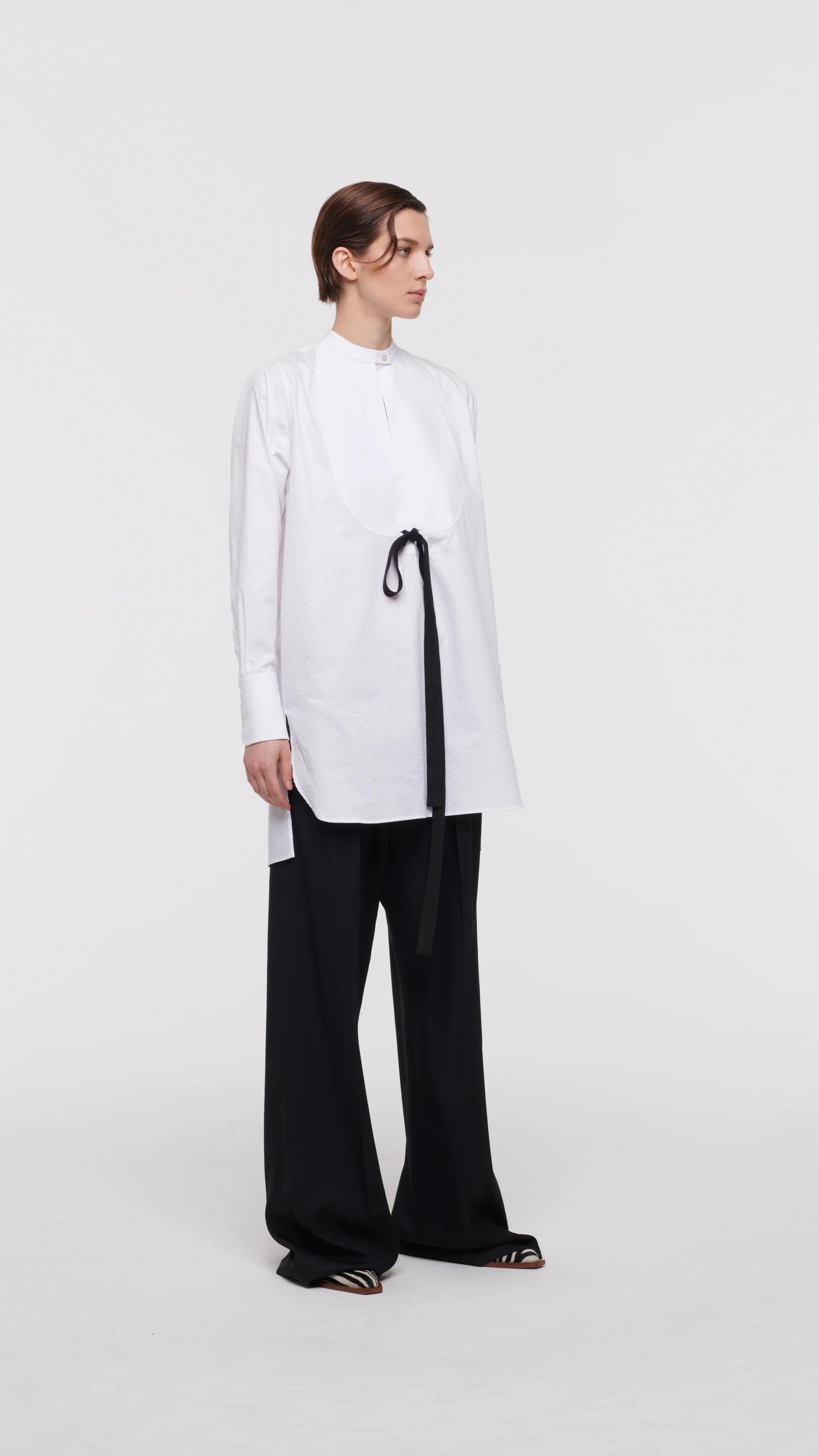 Plan C White Sartorial Poplin Shirt. Classic with a twist long white poplin blouse with front buttons. An added black ribbon detail in the front. Oversized cuffs at the long sleeves. Product photo shown from the side.