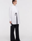 Plan C White Sartorial Poplin Shirt. Classic with a twist long white poplin blouse with front buttons. An added black ribbon detail in the front. Oversized cuffs at the long sleeves. Product photo shown from the side.