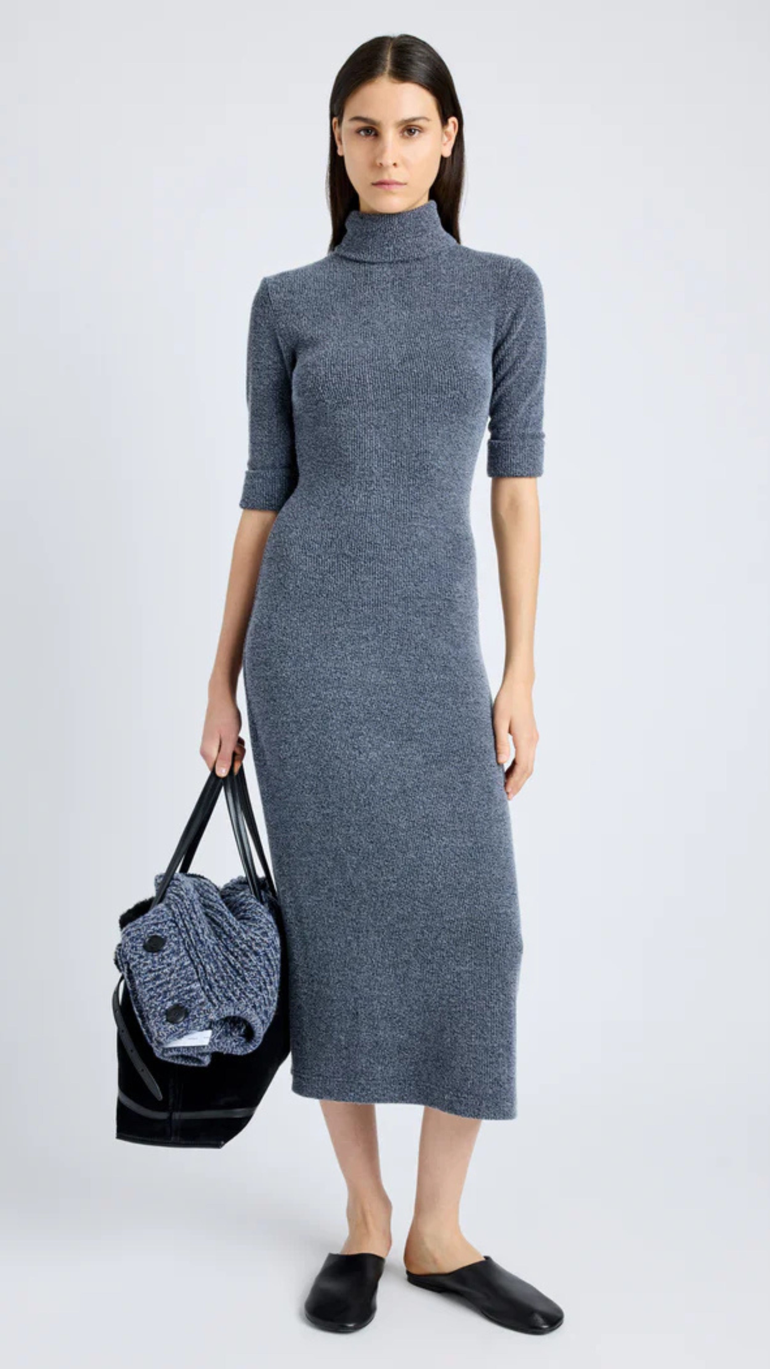 Woman wearing grey blue boucle knit dress with mock turtle neckline, cap sleeves, and midi length, holding a black bag.