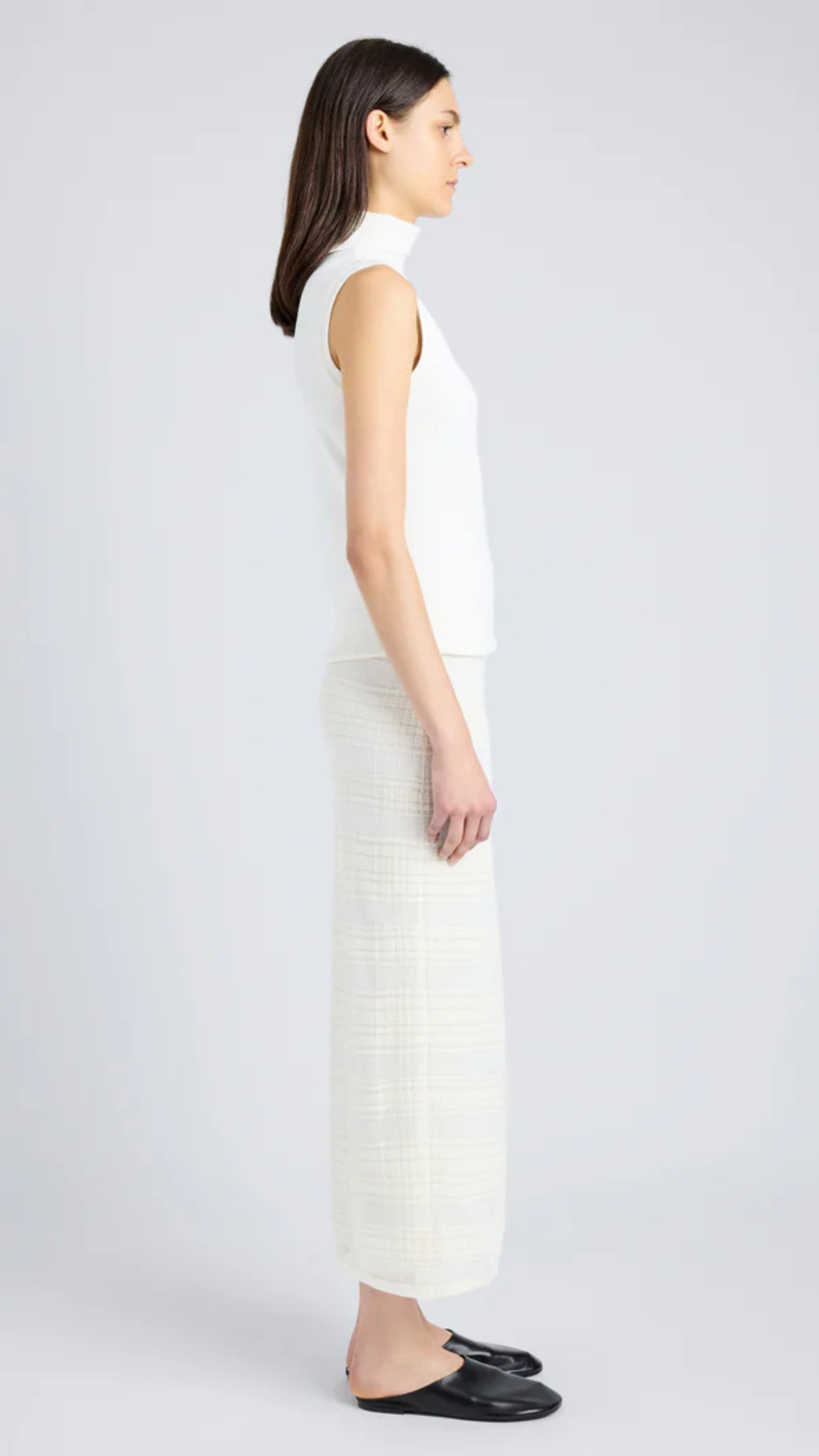 Side view of model wearing a semi-sheer alabaster white plaid knit midi skirt with slip-on design and elastic waist, paired with a white top.