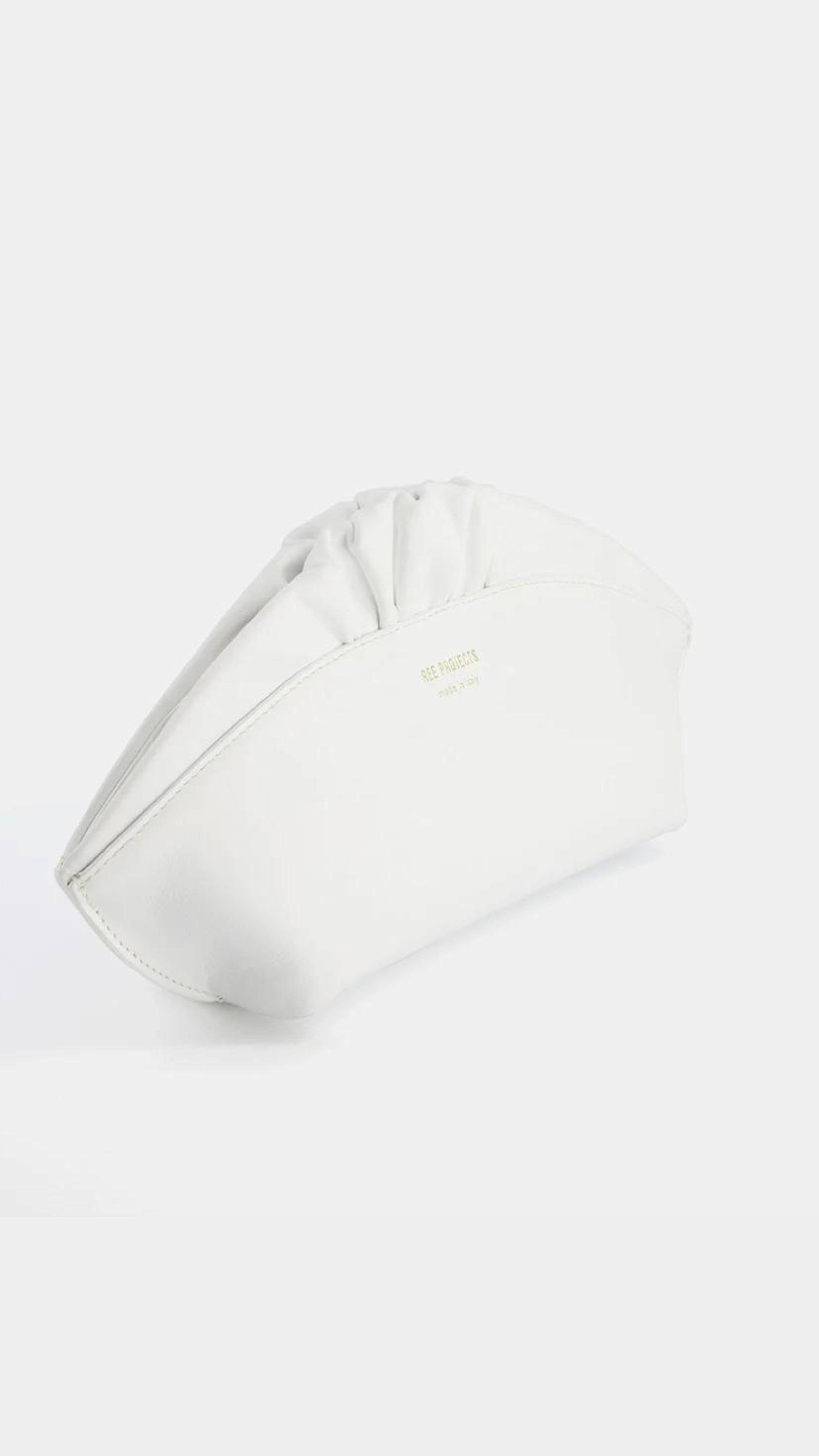 Ree Projects, Ann Baguettina in White. Made in the softest Italian leather this clutch style is perfect for any occasion. It has a slim curved top that is finished with a delicate ruffle and gold hardware. It features a strong magnetic closure and has internal straps to allow it to convert to a shoulder or crossbody bag.  Shown from side angle.