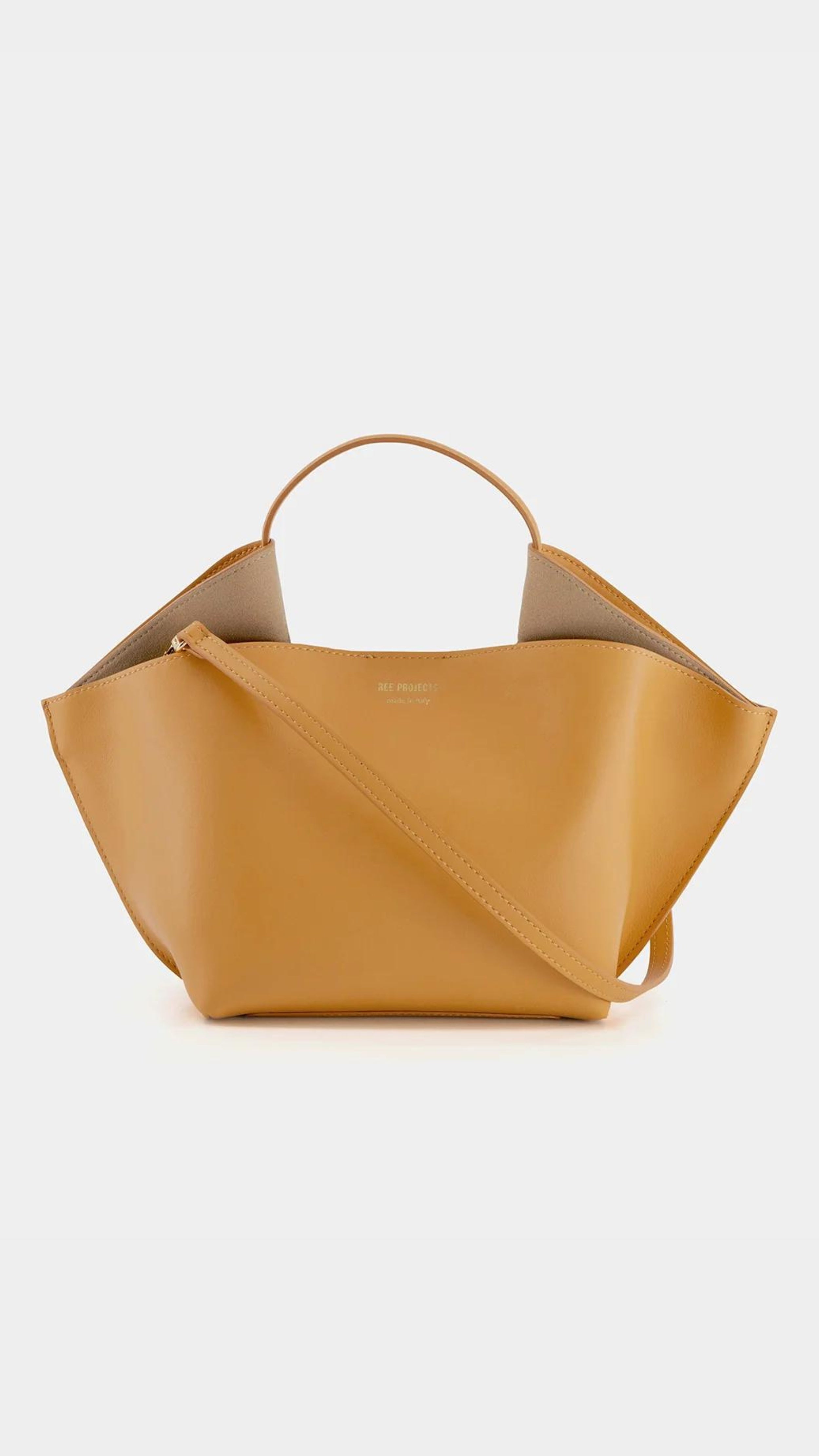 Ree Projects, Anne Tote Mini in Apricot.  An understated elegant every day bag. Crafted from the softest Italian calf leather, this apricot orange  purse features the signature folded &#39;Ree Projects top-line gussets&#39; and a sculptural silhouette. Its mini-sized size easily fits your phone, tablet and other items. This purse can be used with the top handle or as a crossbody. It features interior pockets and a magnet snap closure.  Shown from the back with crossbody strap.