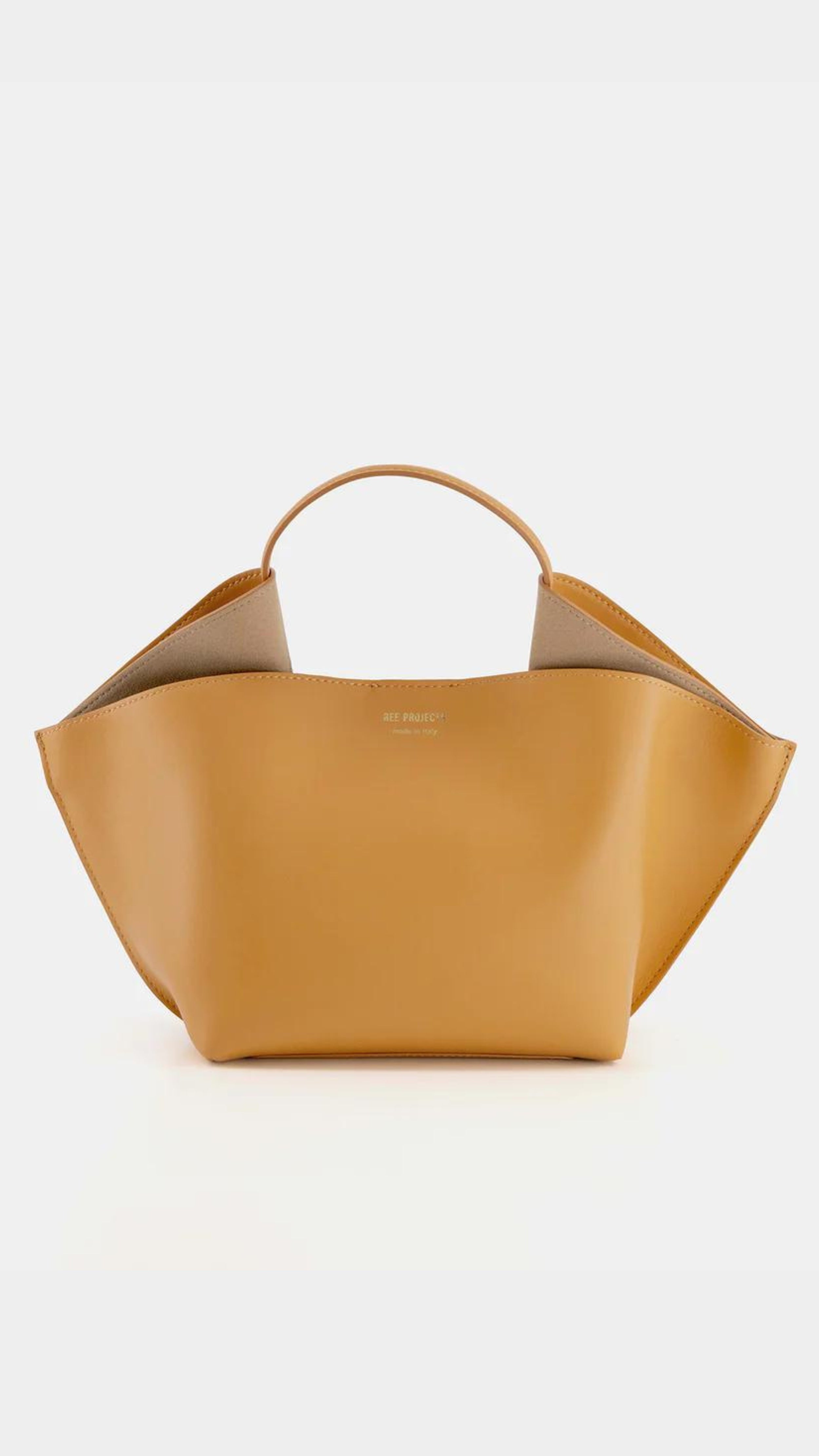Ree Projects, Anne Tote Mini in Apricot.  An understated elegant every day bag. Crafted from the softest Italian calf leather, this apricot orange  purse features the signature folded 'Ree Projects top-line gussets' and a sculptural silhouette. Its mini-sized size easily fits your phone, tablet and other items. This purse can be used with the top handle or as a crossbody. It features interior pockets and a magnet snap closure.  Shown from the front view.