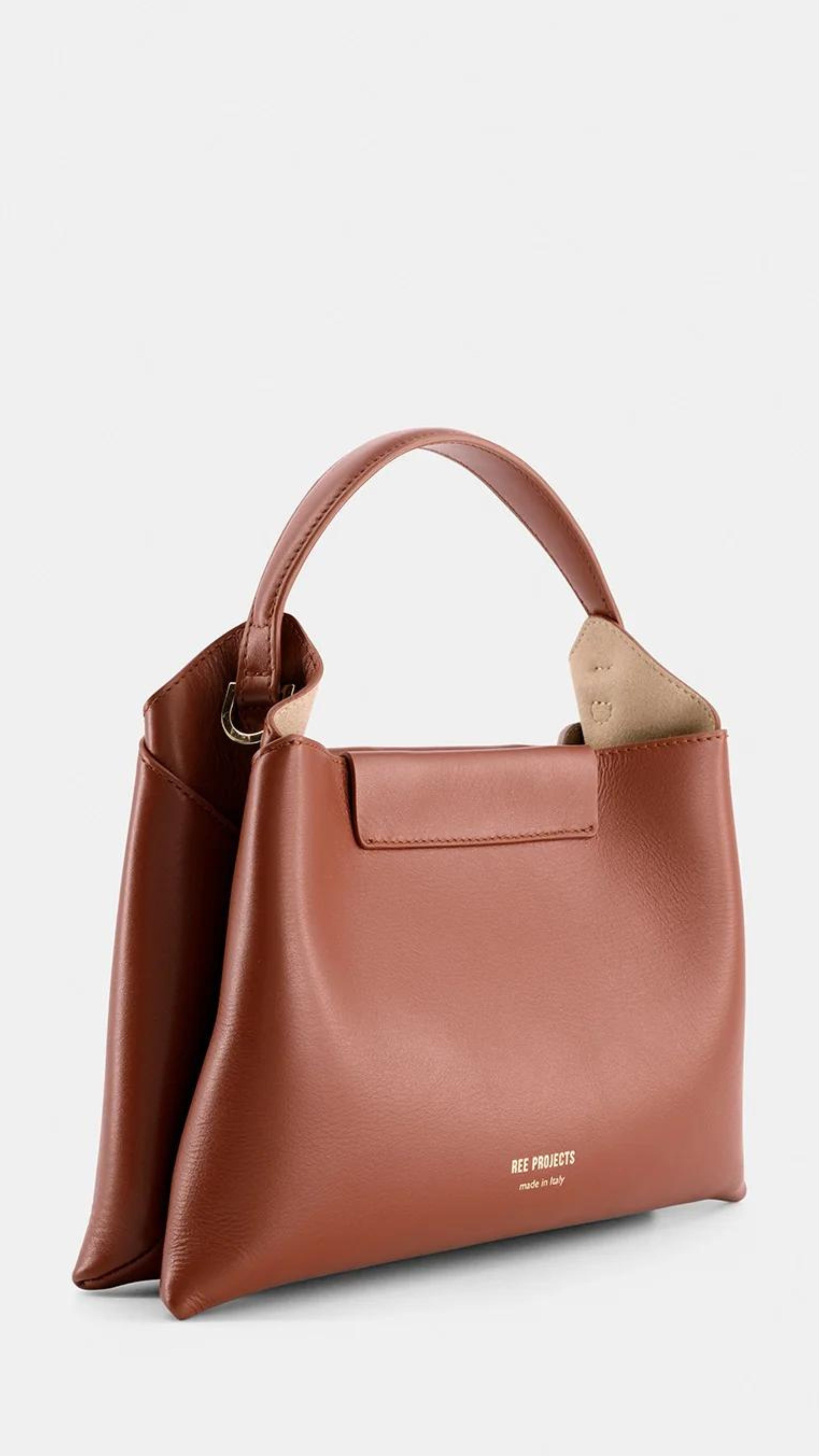 Ree Projects Elize Medium Bag in Cognac Brown. Made in italy and sustainable. Squared shape, it features a magnetic button closure and signature gussets. The purse has a spacious interior, padded top handle, and optional shoulder strap. Shown from the front side view.