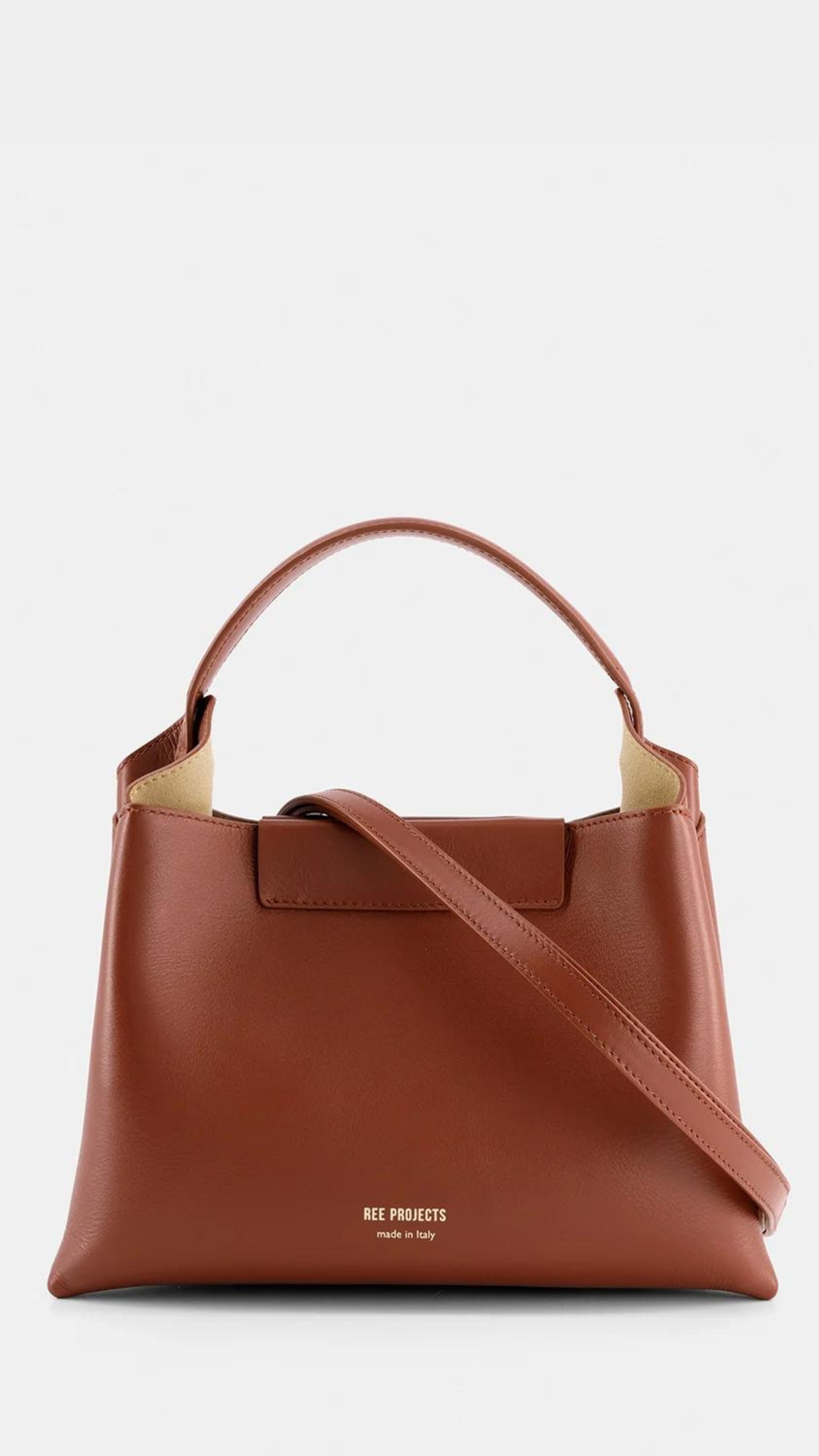Ree Projects Elize Medium Bag in Cognac Brown. Made in italy and sustainable. Squared shape, it features a magnetic button closure and signature gussets. The purse has a spacious interior, padded top handle, and optional shoulder strap. Shown from the front view.