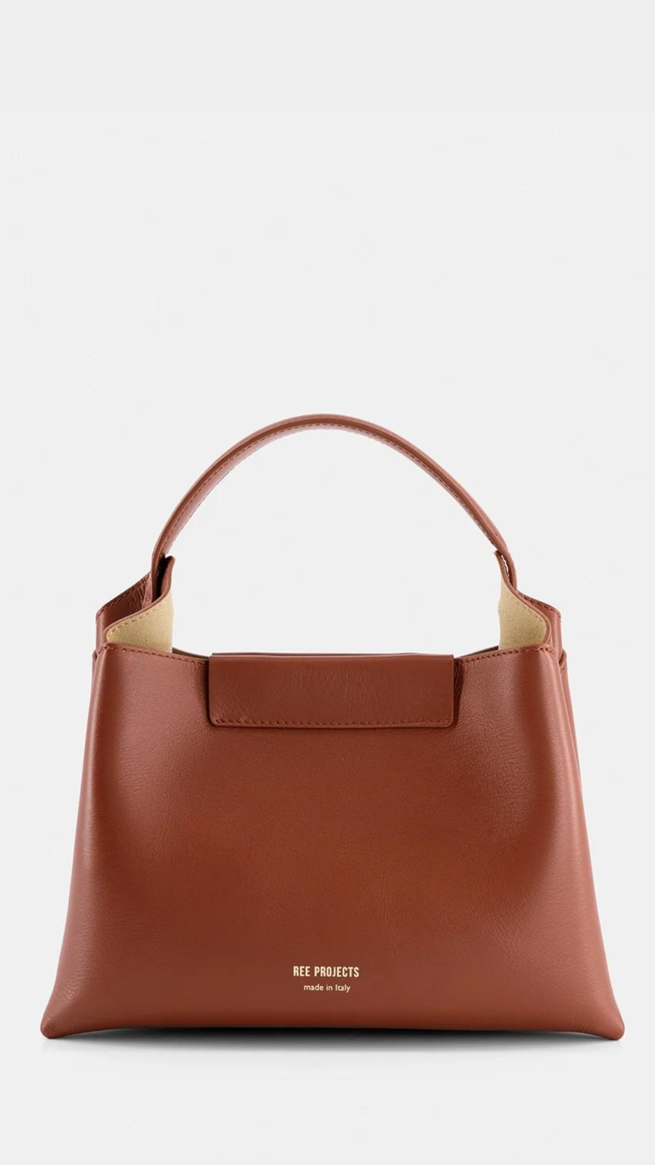 Ree Projects Elize Medium Bag in Cognac Brown. Made in italy and sustainable. Squared shape, it features a magnetic button closure and signature gussets. The purse has a spacious interior, padded top handle, and optional shoulder strap. Shown from the back view.