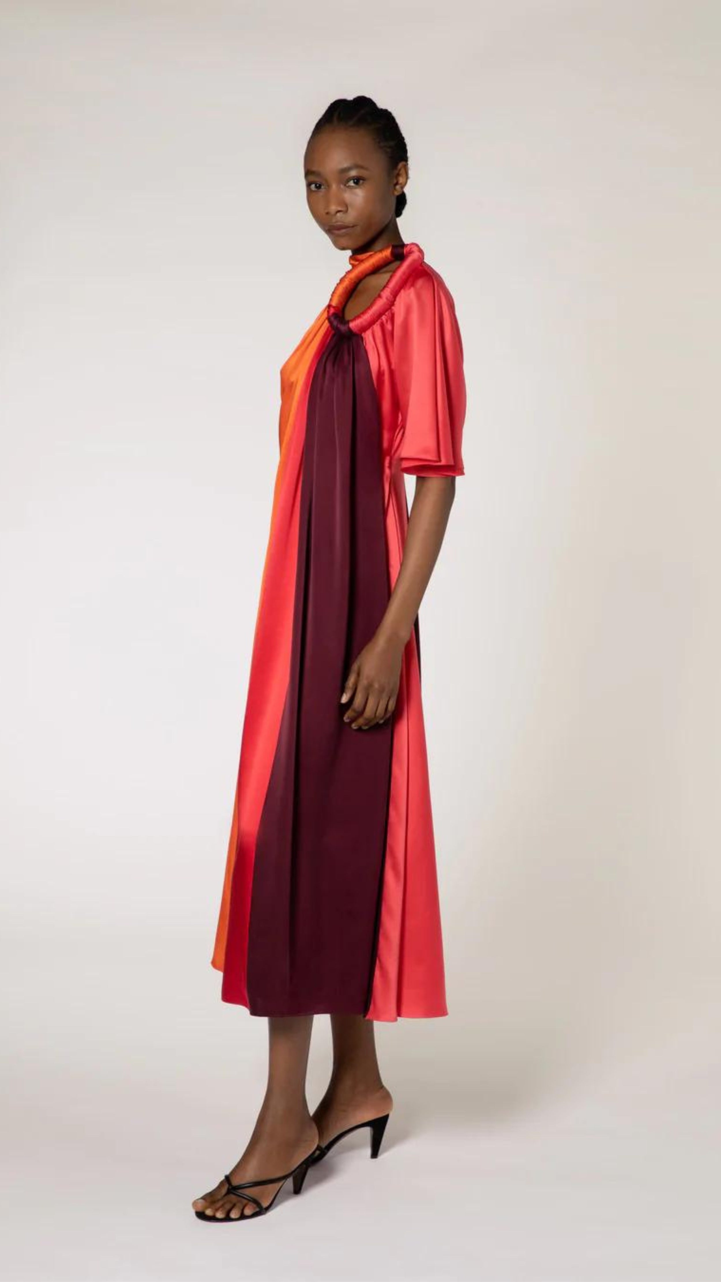 Roksanda Fiamma Dress. Silk midi dress in vertical panels of orange, rose red and burgundy. Features an architectural oval cut out at the top right front. Shown on model facing side.