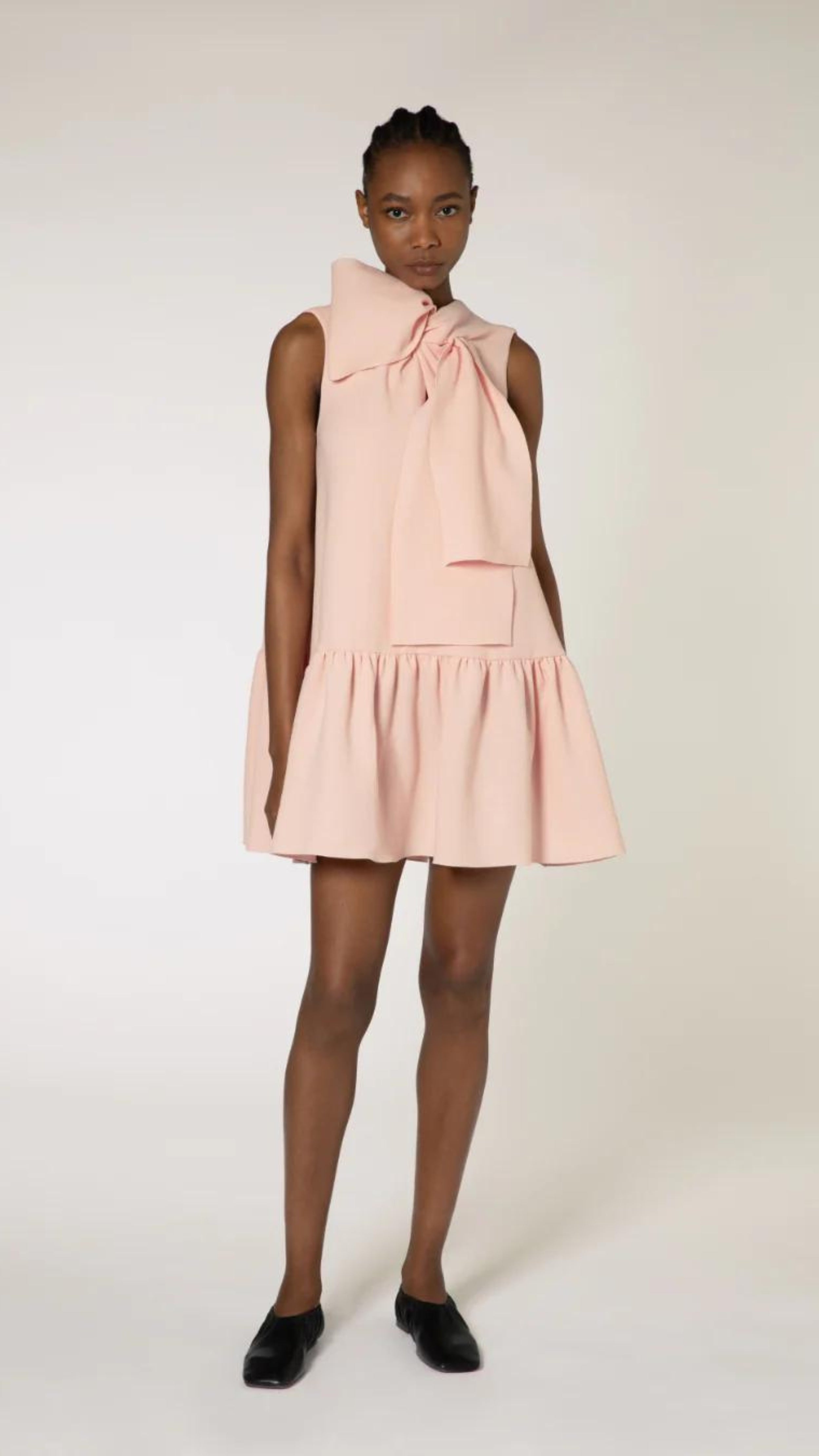 Roksanda Rosalina Dress. Soft pink with fun oversize twisted bow at neckline. Mini dress length with a dropped waist and pleated skirt panel. Shown on model facing front.