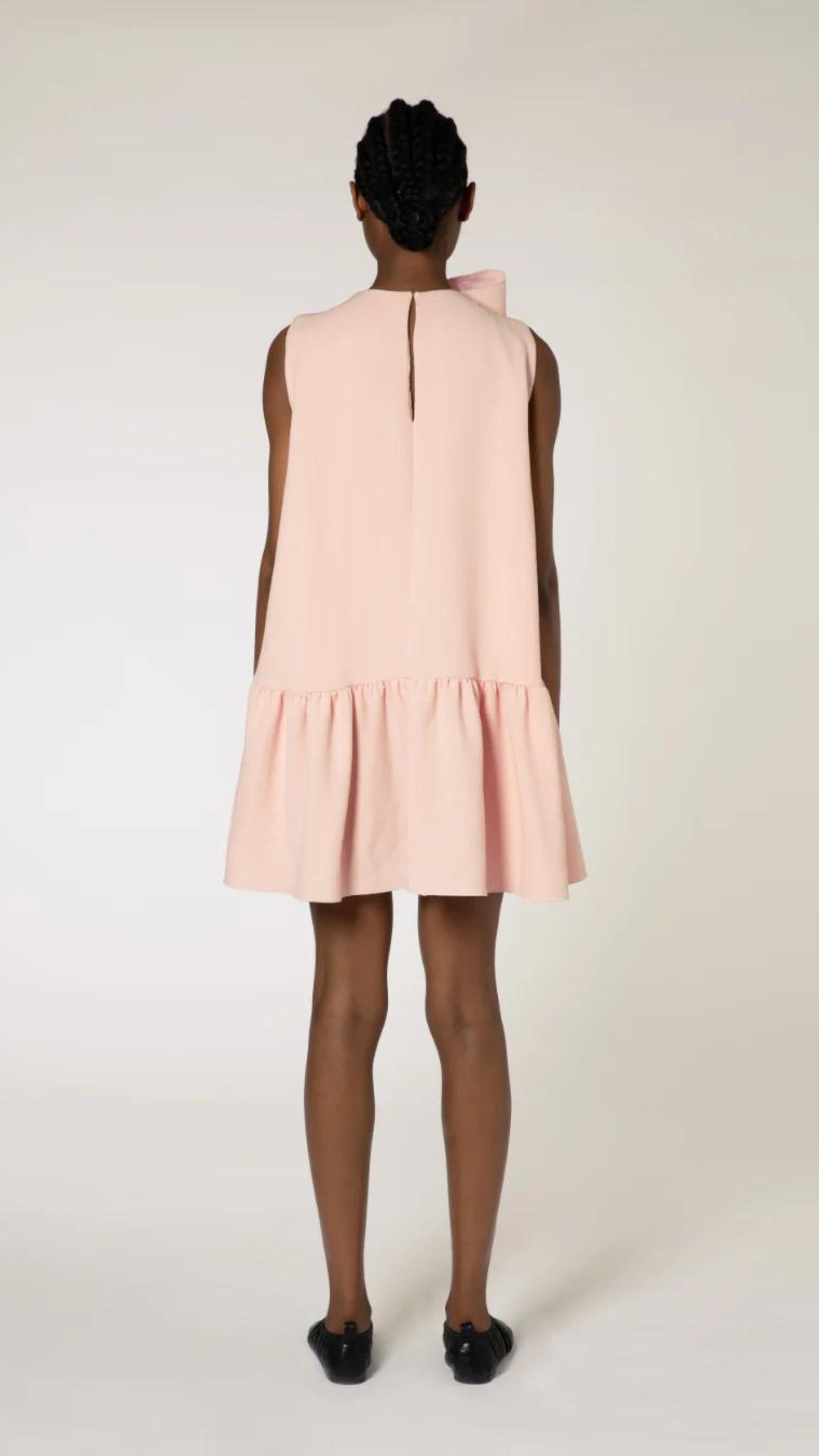 Roksanda Rosalina Dress. Soft pink with fun oversize twisted bow at neckline. Mini dress length with a dropped waist and pleated skirt panel. Shown on model facing the back.