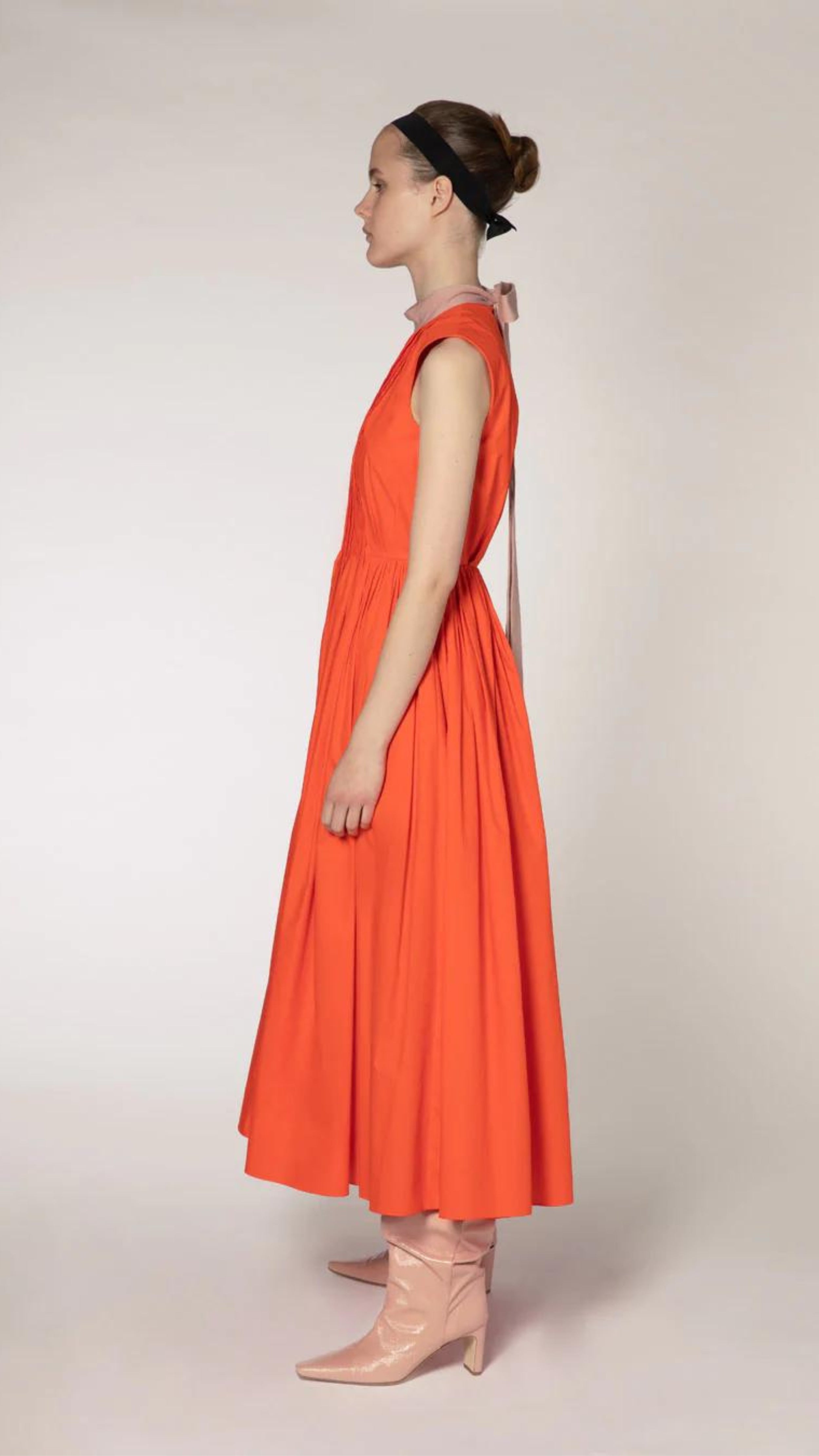 Roksanda The Prinesa Dress. Classic elegant orange cotton dress with a rounded neckline, fitted waist and pleated midi length skirt. Highlighted with a pale pink ribbon collar that ties at the nape of the neck. Shown on model facing side.