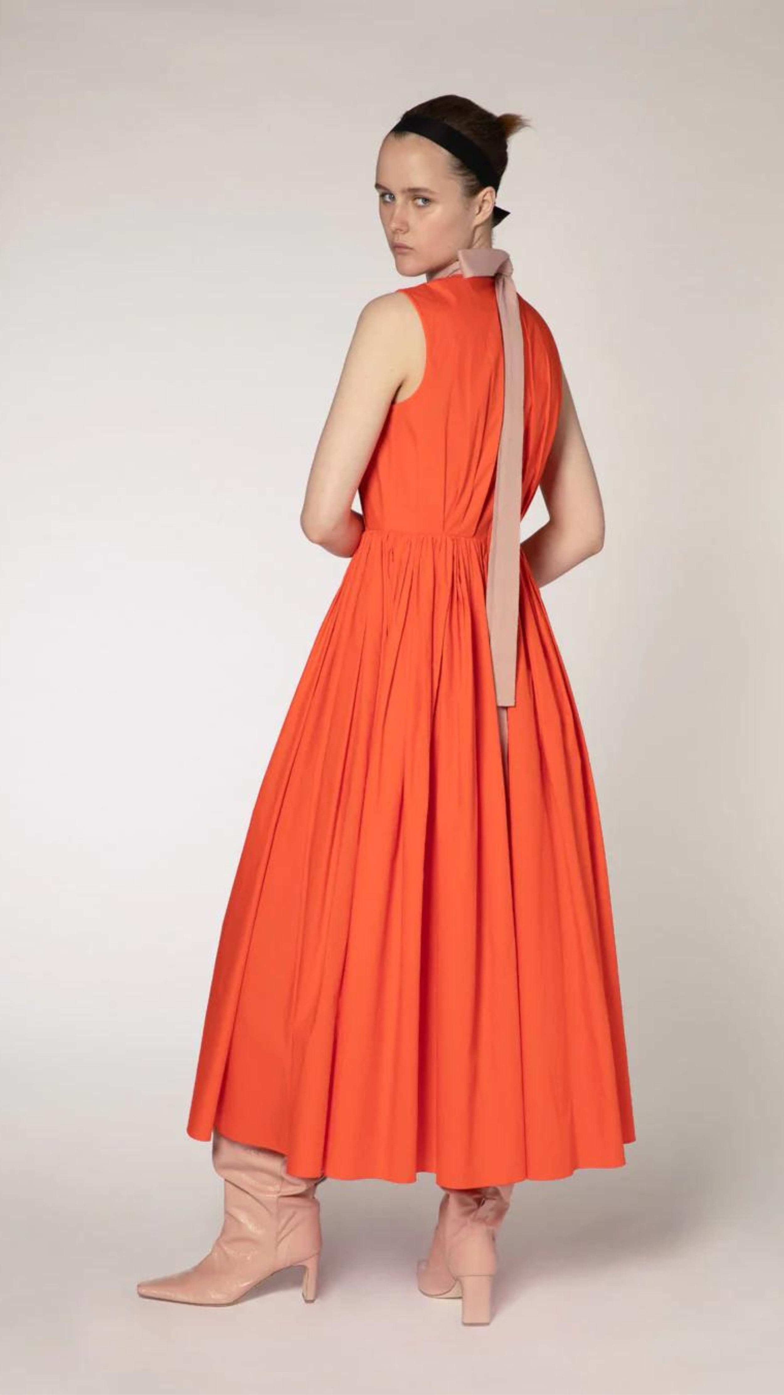 Roksanda The Prinesa Dress. Classic elegant orange cotton dress with a rounded neckline, fitted waist and pleated midi length skirt. Highlighted with a pale pink ribbon collar that ties at the nape of the neck. Shown on model facing back.