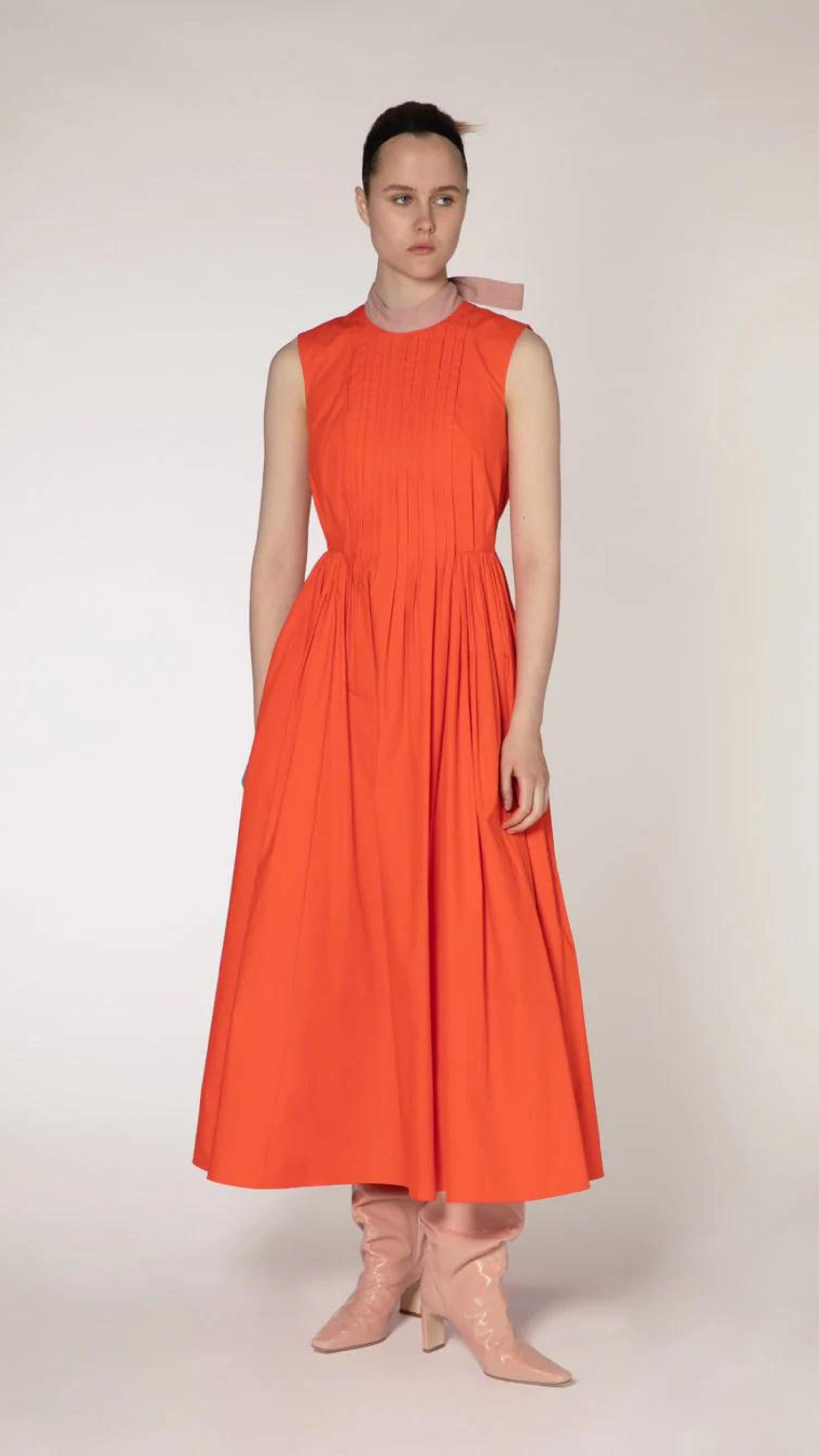 Roksanda The Prinesa Dress. Classic elegant orange cotton dress with a rounded neckline, fitted waist and pleated midi length skirt. Highlighted with a pale pink ribbon collar that ties at the nape of the neck. Shown on model facing front.